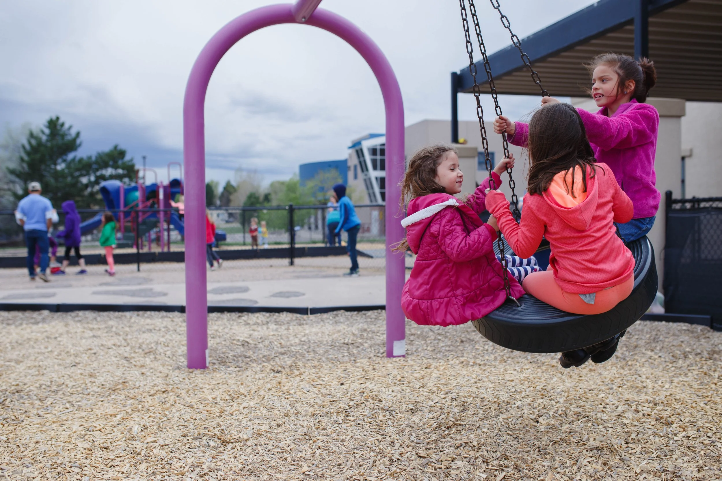 Three young girls playing on a tire swing at a playground, with other children and adults in the background under an overcast sky.