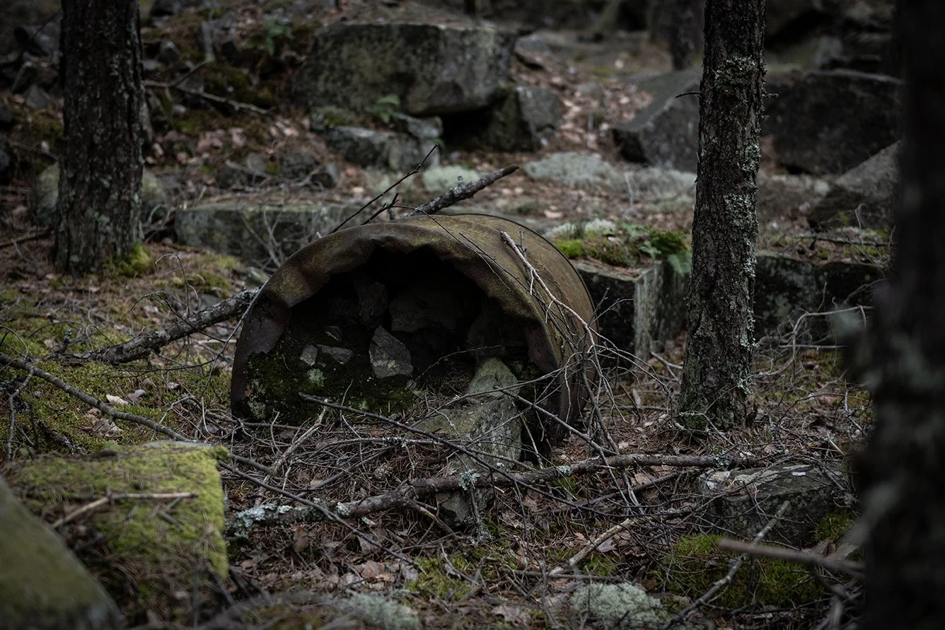 A rusty, cylindrical metal pipe lying on the forest ground surrounded by rocks, fallen leaves, twigs, and moss, with trees in the background.