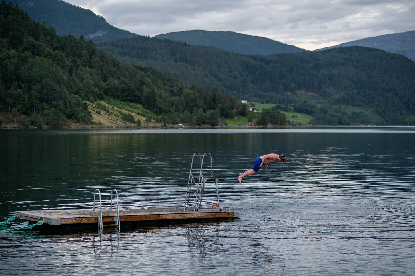 A person is mid-air jumping off a dock into a lake surrounded by green mountains.