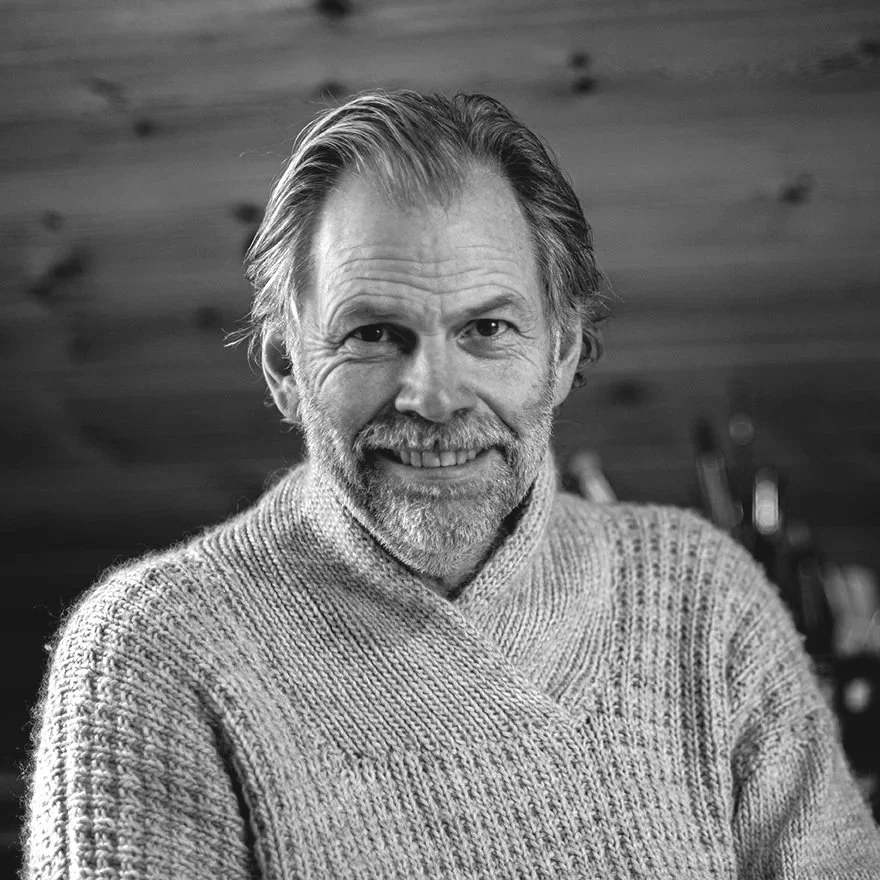 Black and white photo of an older man with a beard and mustache, smiling, wearing a knit sweater, with a wooden wall in the background.