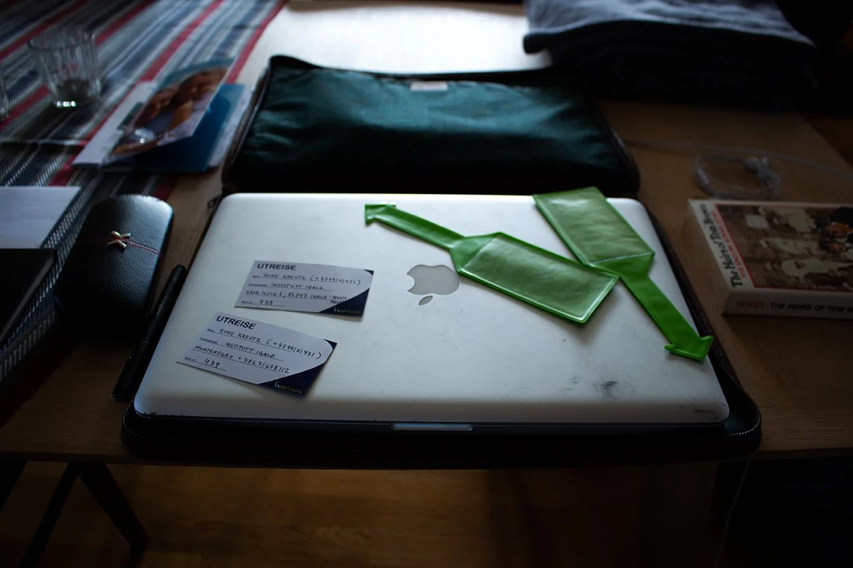 A closed silver Apple MacBook on a wooden desk with two green plastic bags and two boarding passes placed on top. The desk has various items including a black case, a pen, magazines, and a pair of glasses.