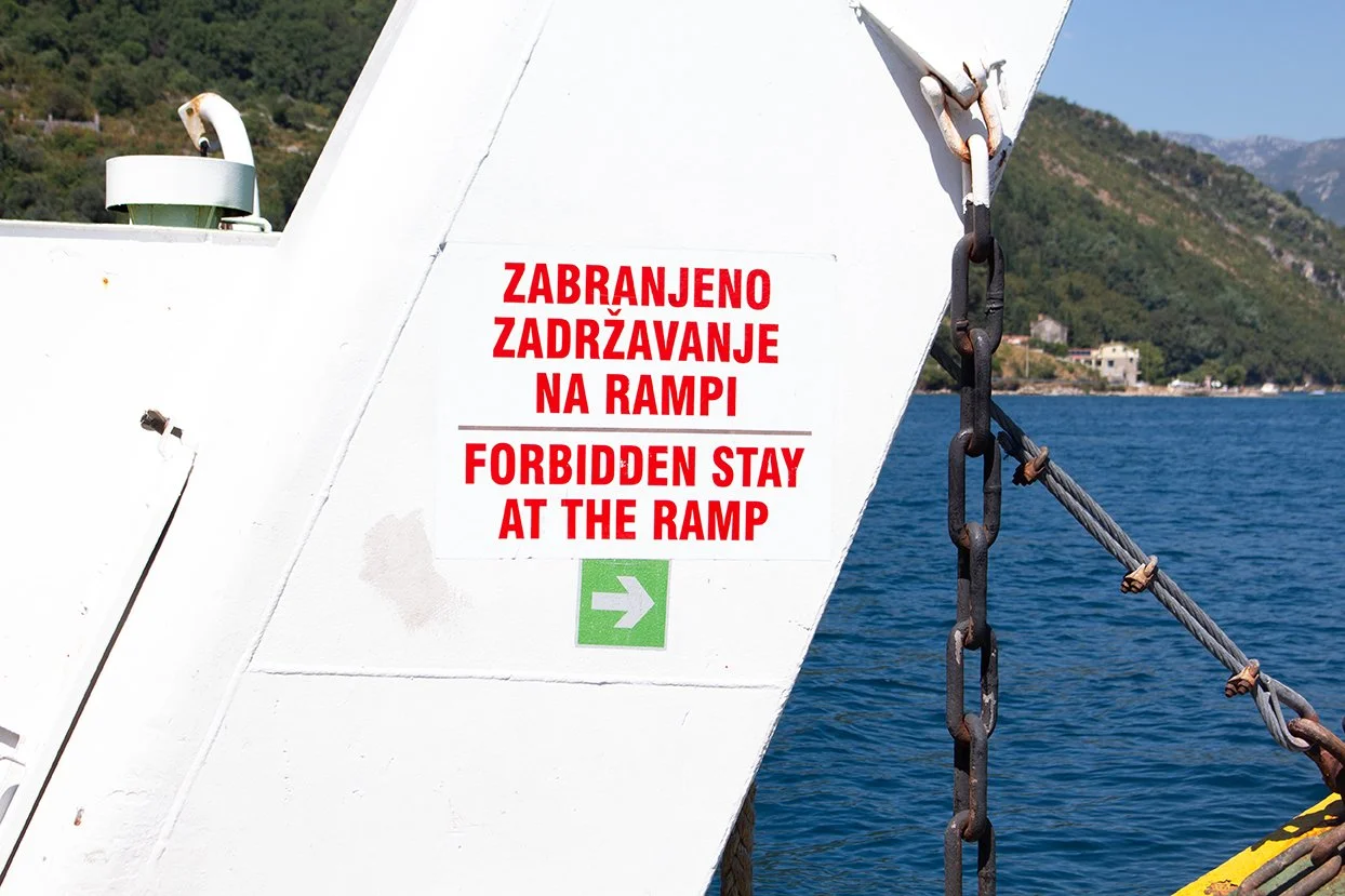 A sign on a ship's white metal structure with text in Croatian and English that reads "Forbidden stay at the ramp". There is a green arrow pointing right, and the background features a body of water and hilly terrain with buildings.