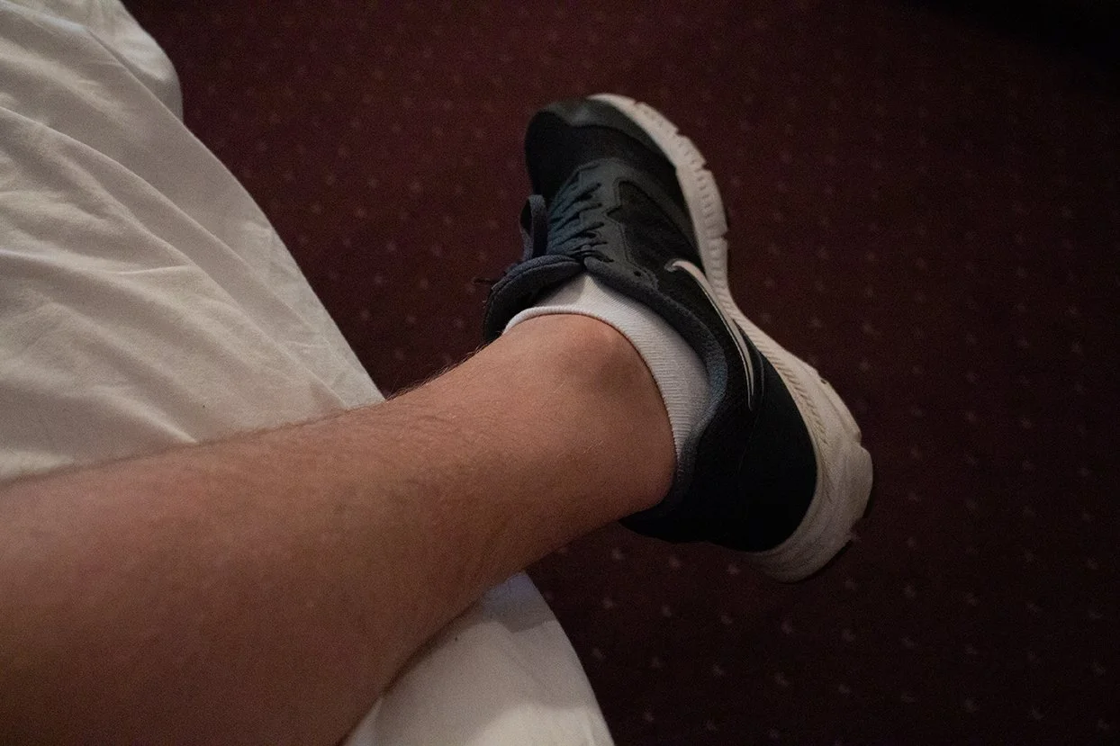 A person's leg and foot wearing a black and white sneaker and white sock, resting on a white bed beside a red carpeted floor.