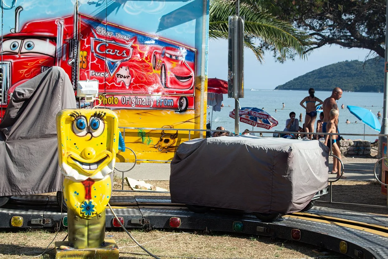 A beachside scene with a Spongebob Squarepants-themed inflatable figure in the foreground. In the background, there are people swimming and relaxing at the beach, with umbrellas and palm trees, and mountains visible across the ocean.