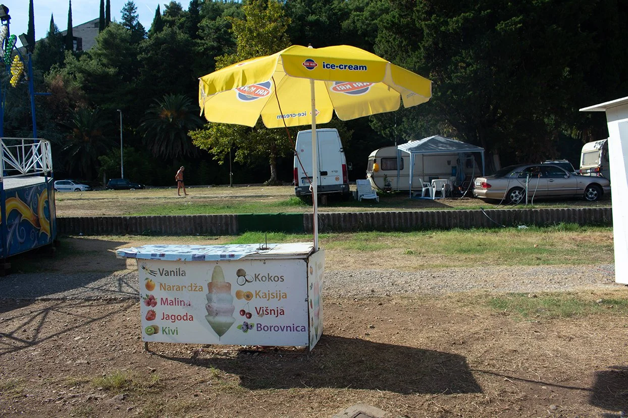 A small ice cream stand with a yellow umbrella, displaying flavors such as vanilla, orange, raspberry, strawberry, kiwi, coconut, cashew, cherry, and blueberry. Behind the stand, there are cars, a trailer, a canopy tent, and a person walking in a gra