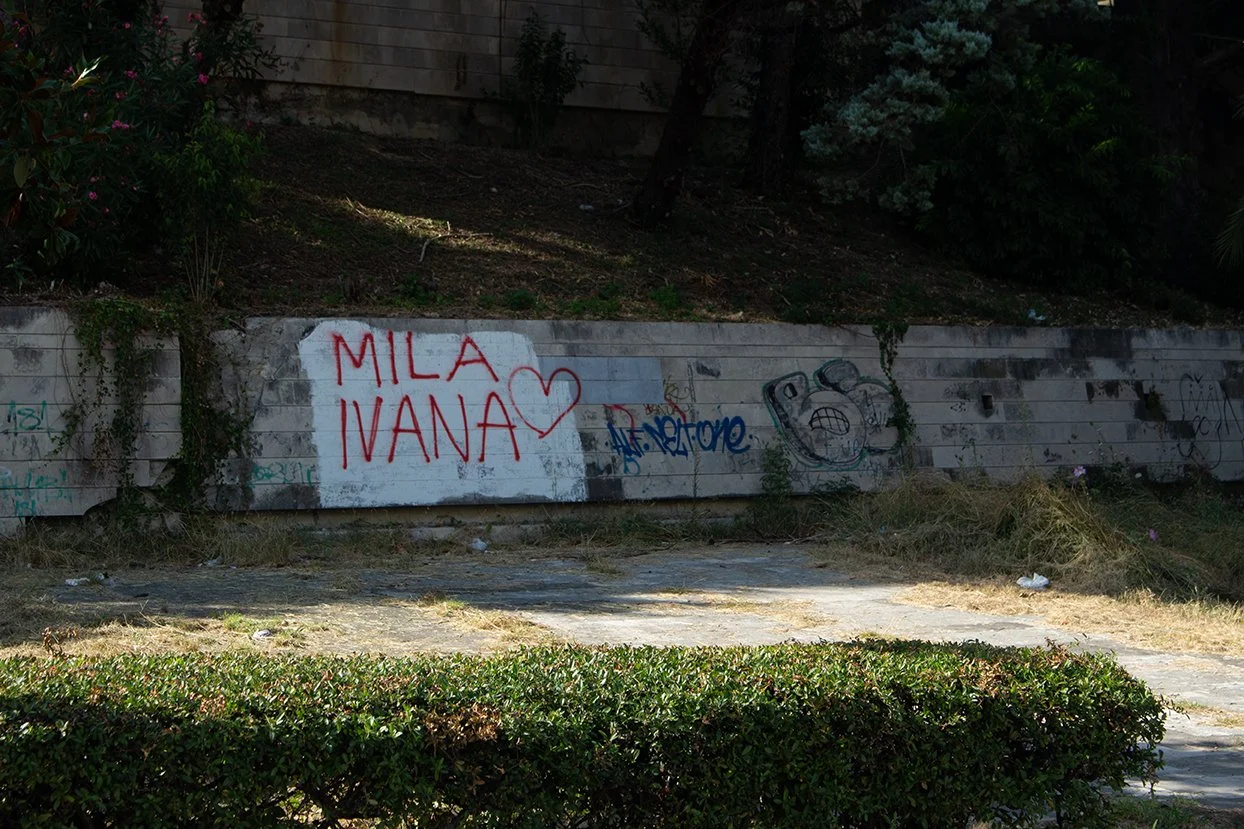 Graffiti on a concrete wall with the words 'MILA IVANA' and a heart, along with other tags and a cartoonish face.