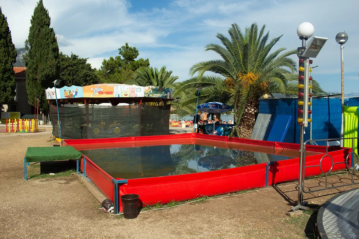An outdoor carnival game features a water-filled red rectangular tank with a small green platform on the side, a metal pole with colorful lights, and a canopy with cartoon characters in the background. There are palm trees, a blue sky, and people sit