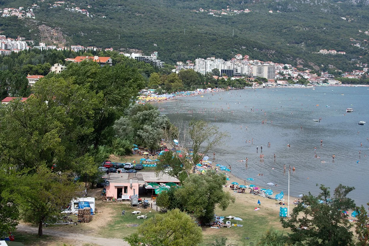 A scenic view of a beach with many people swimming and sunbathing. There are colorful umbrellas and boats on the water, with a backdrop of green hills and a cityscape with high-rise buildings.