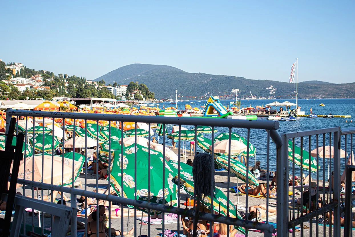 Beach scene with umbrellas, people sunbathing, and a view of the water and mountains in the background.