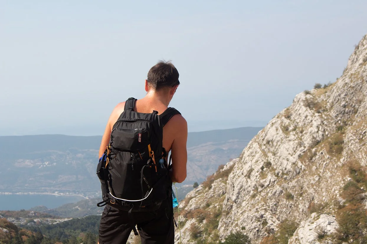 A man with a backpack hiking near rocky cliffs with a mountain and water in the background.