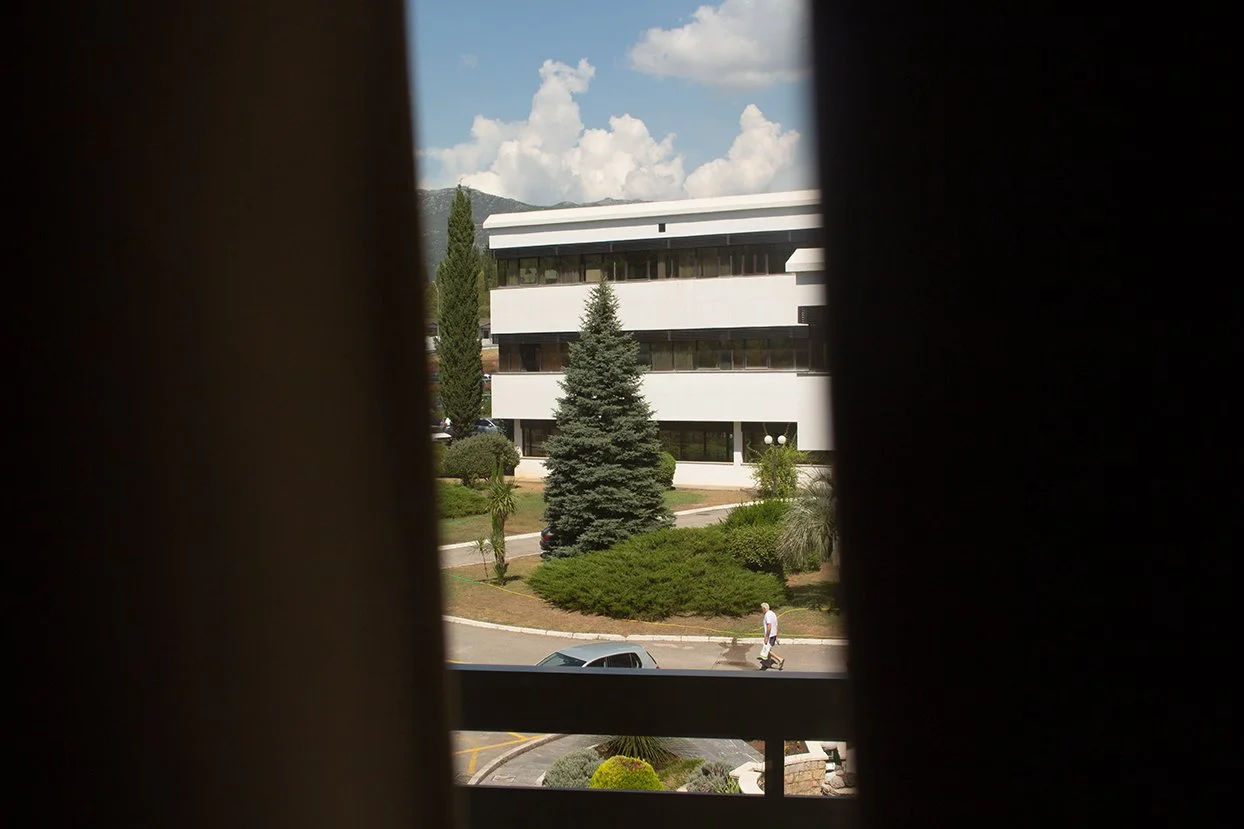 Photo taken through a window with dark curtains, showing a street scene outside. There is a large modern white building with multiple floors, trees, bushes, and a cloudy sky. A person is walking on the sidewalk pulling a small suitcase or bag.