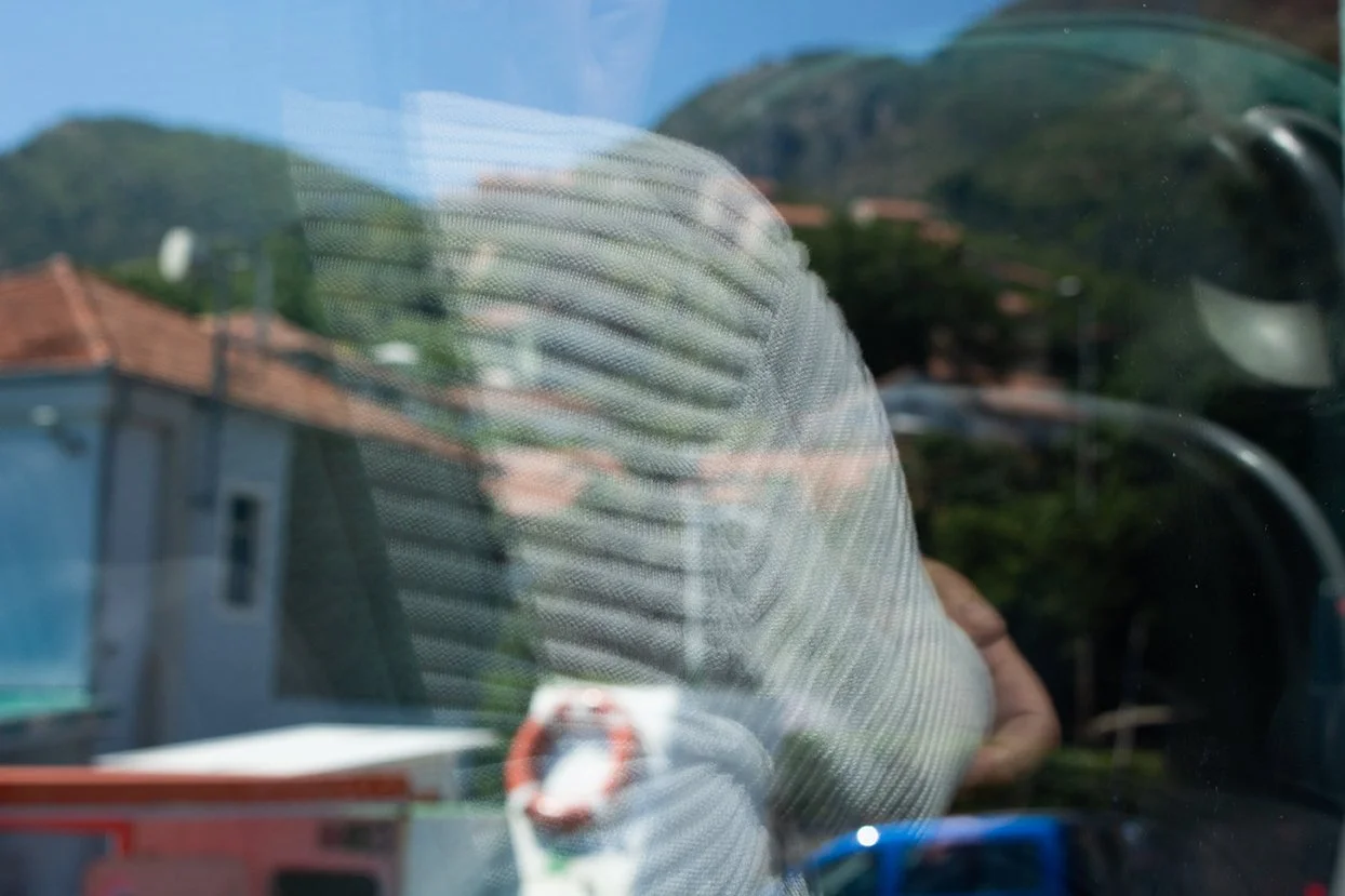 Reflection of a person wearing a striped hoodie taking a photo through a glass window during daytime with houses, trees, and hills in the background.