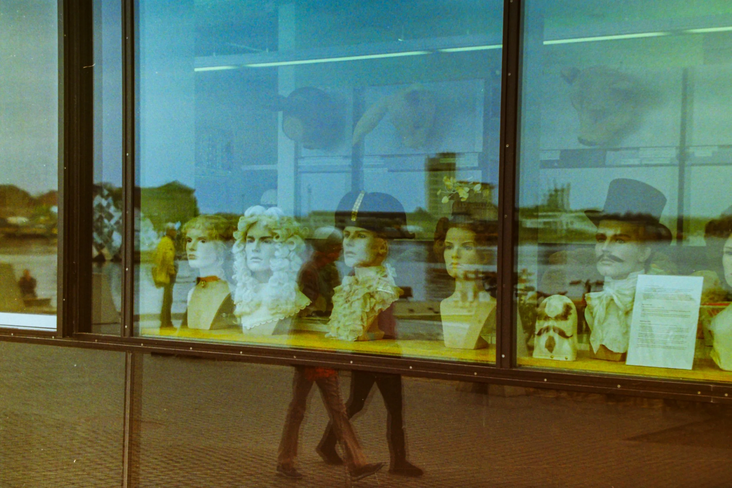 Museum window display featuring various mannequins with historical costumes and wigs, reflections of people walking by and landscape outside.