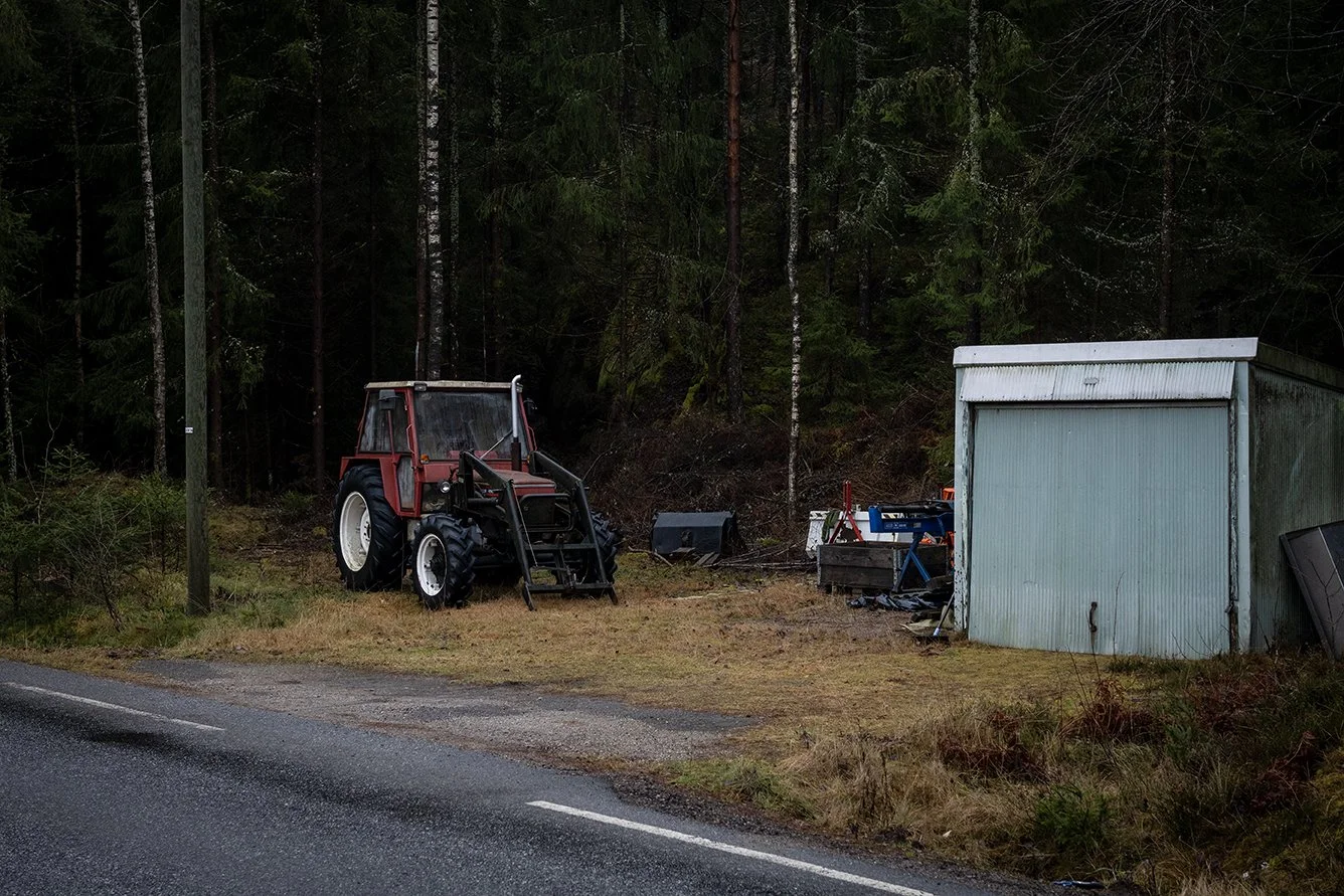 A red tractor parked on a grassy area beside a road, next to a small weathered metal shed and some outdoor equipment, with a forested background.