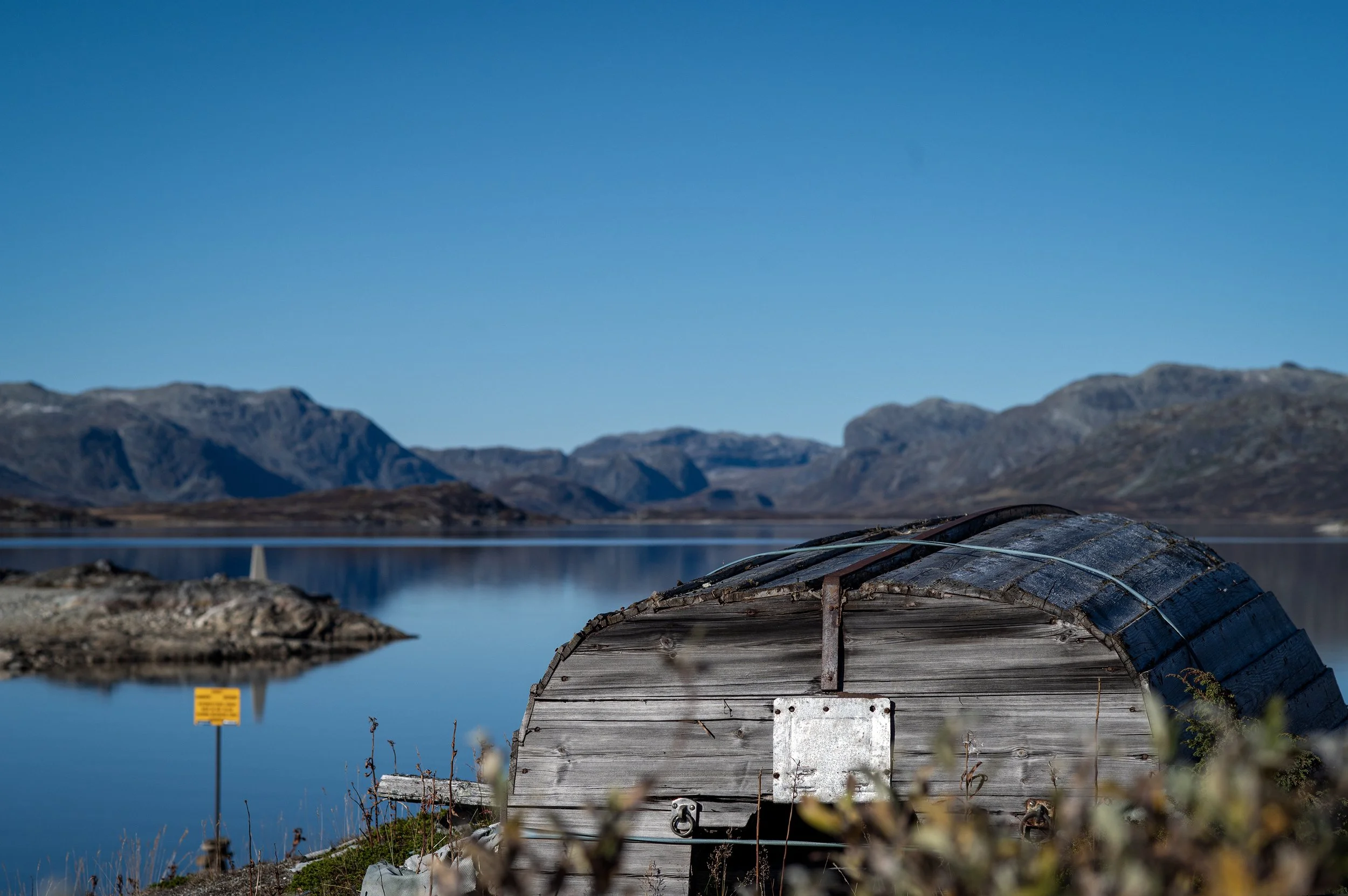 A rustic wooden boat with a curved roof on the shore of a calm lake, surrounded by mountainous terrain under a clear blue sky.