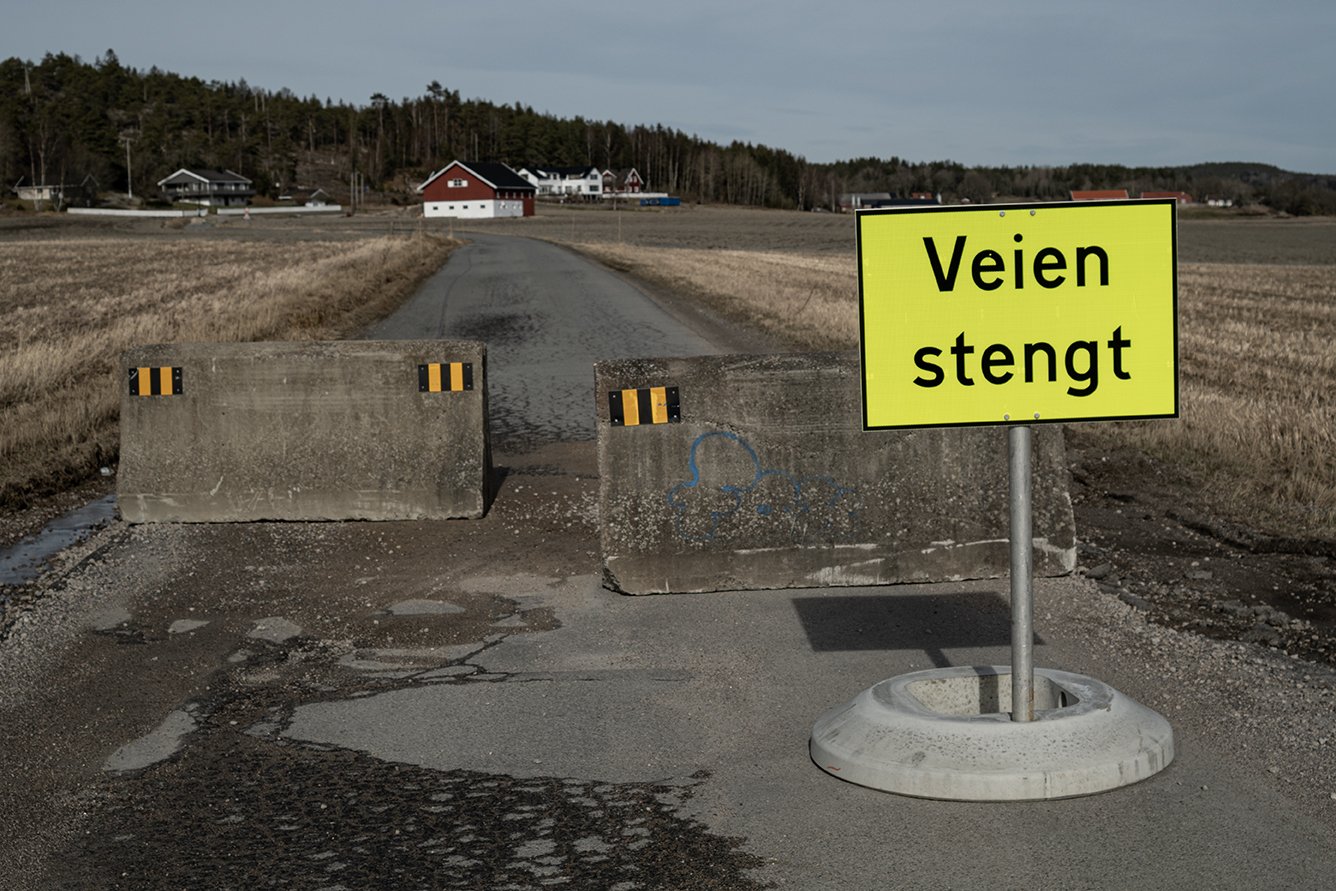 A gravel road is blocked by concrete barriers and a bright yellow sign that says 'Veien stengt' in Norwegian, which means 'Road closed.' In the background, there are houses and trees under a cloudy sky.