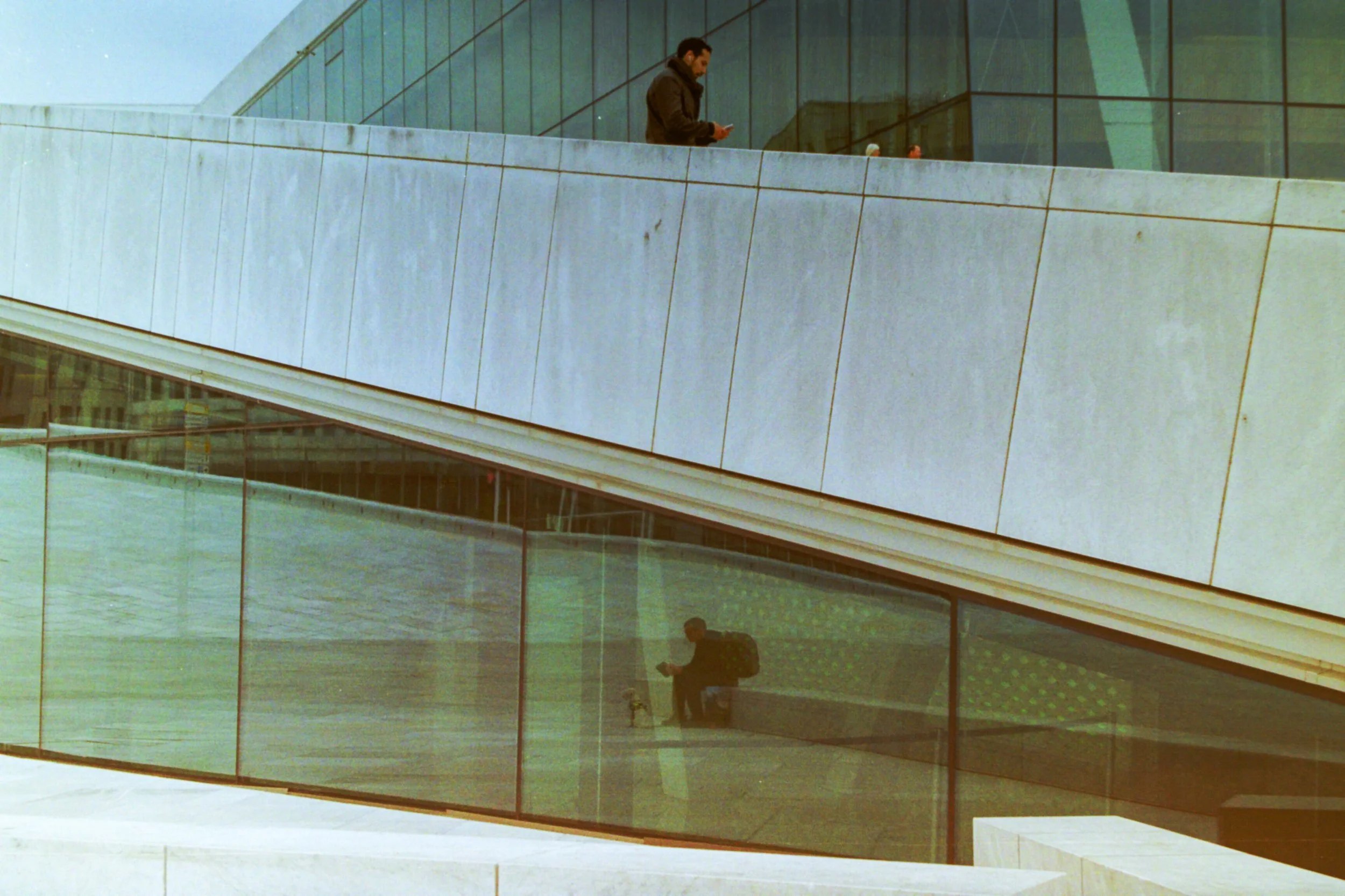 Two men are on different levels of a modern building with glass and concrete architecture. One man is on the upper level looking at his phone, and the other is sitting below, also looking at his phone.