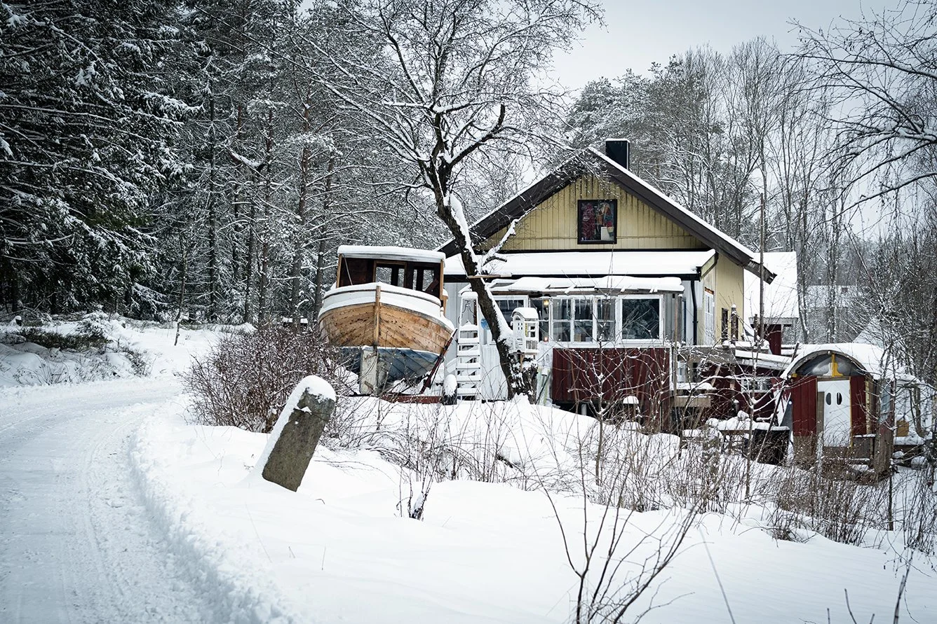 Snow-covered house surrounded by trees, with a boat on a trailer and a snow-covered driveway.