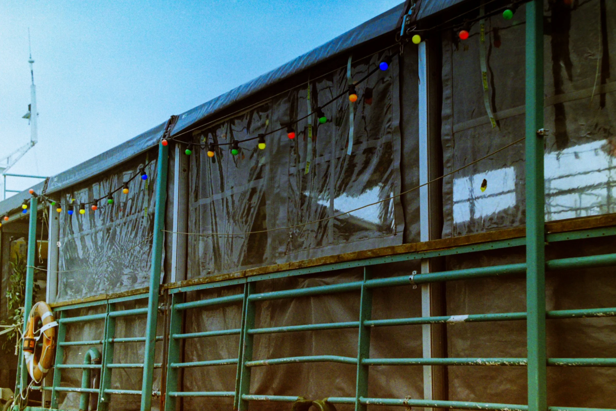Outdoor structure with transparent plastic walls, green metal railings, and a string of colorful lights. A life preserver is hanging on the left side.