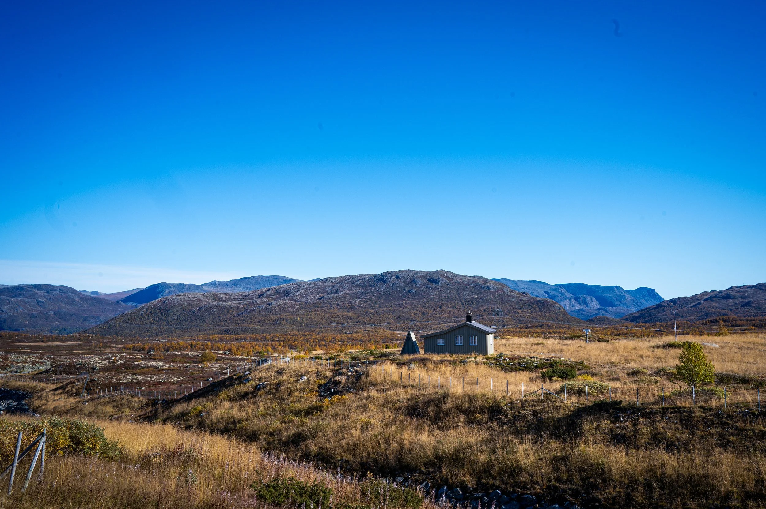 Scenic landscape with a small house in a vast open field, mountains in the background, and clear blue sky.