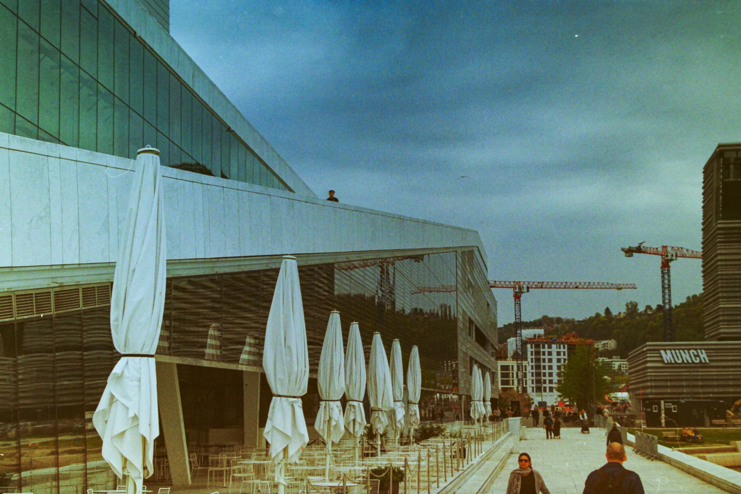 Modern building with glass exterior, outdoor seating with white umbrellas, construction cranes in the background, people walking on sidewalk.