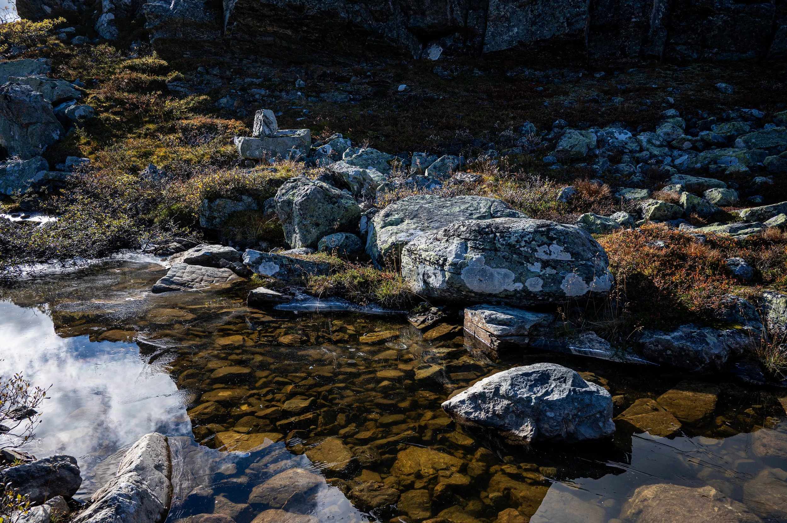 A mountain stream with rocks and boulders, surrounded by vegetation, with reflecting water and a dark background of mountain terrain.