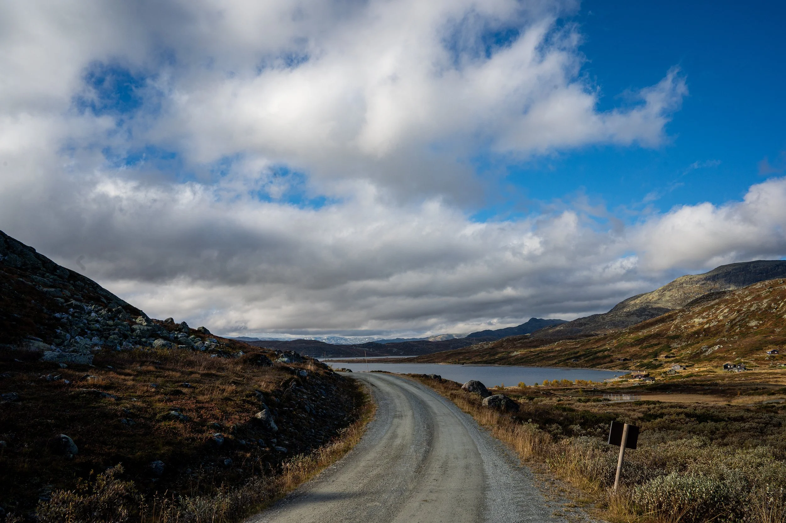 Unpaved mountain road along a lake, with hills and mountains in the distance, partly cloudy sky overhead.