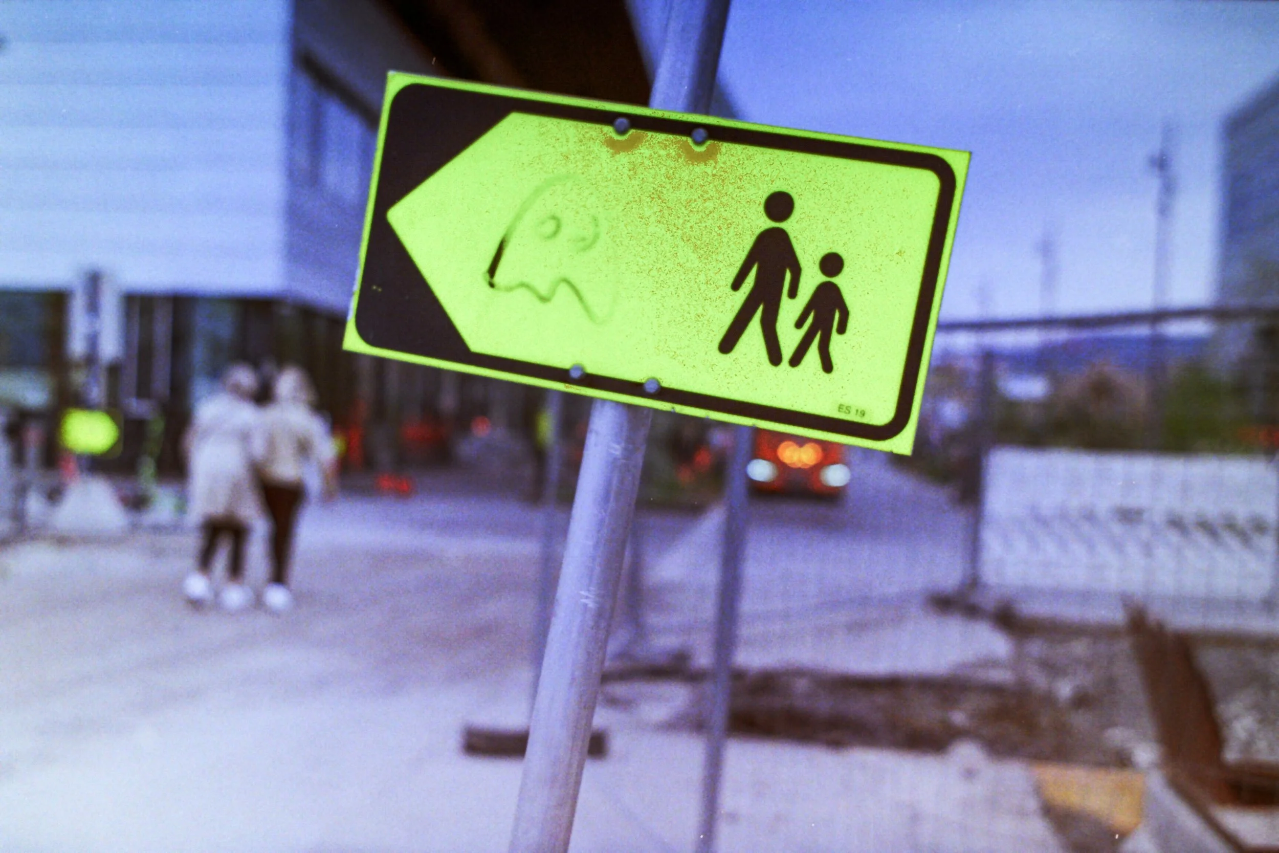 A bright yellow pedestrian crossing sign with a black border, showing two people walking, and a stylized face of a monster or creature in the background. The scene is outdoors with a city street in the background, including a crosswalk, two blurred p