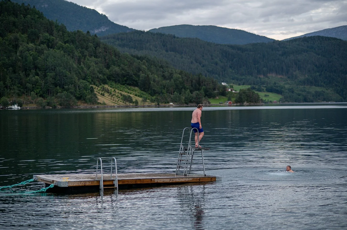 A man in blue swim trunks walking on a small dock with stairs, while another person swims nearby in a lake surrounded by green hills and mountains.