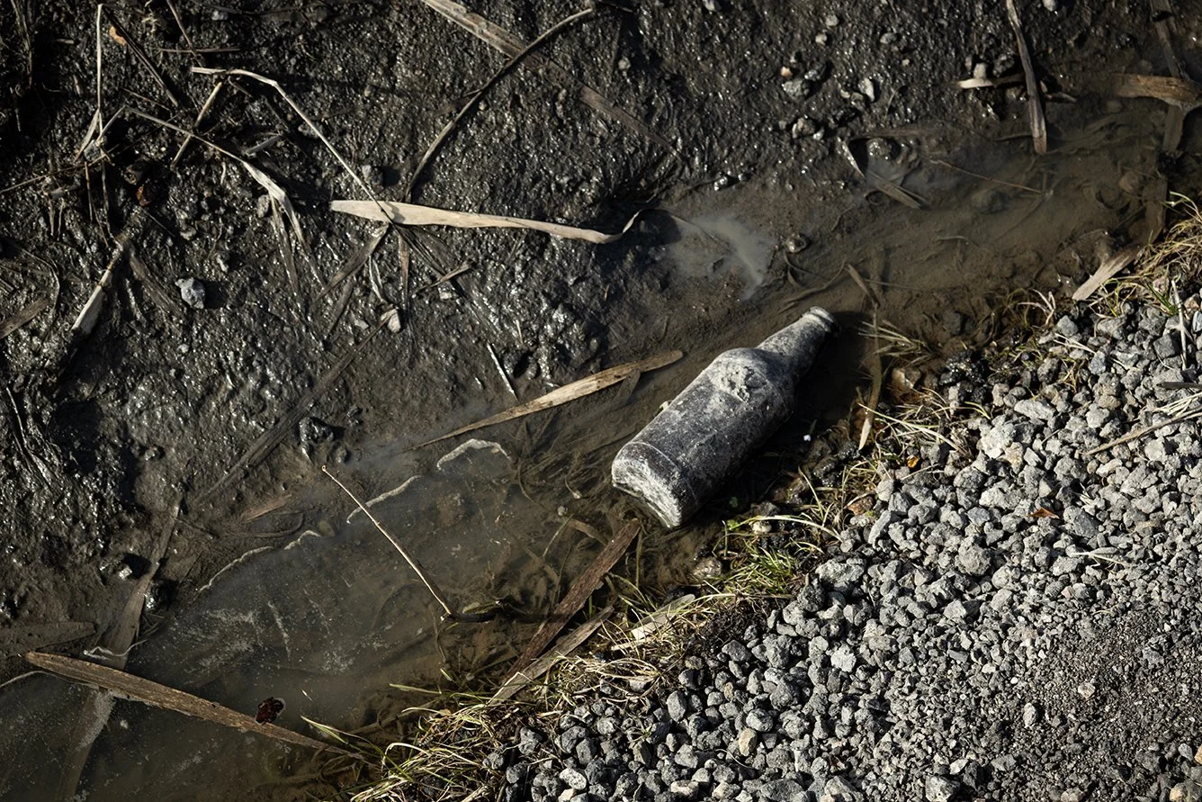 An abandoned, dusty glass bottle lying in muddy water beside gravel and dry grass.