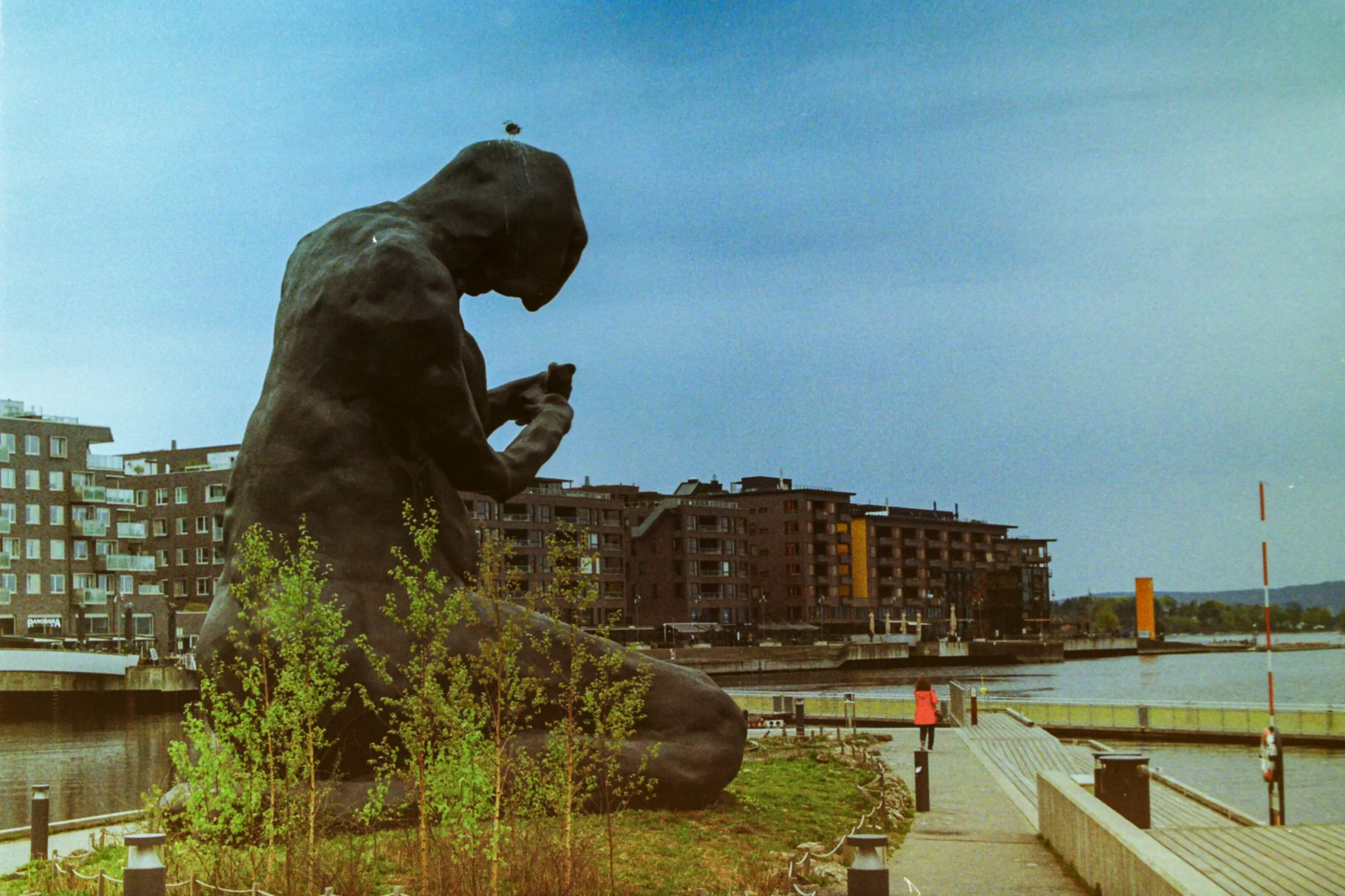 A large bronze statue of a seated man holding an object, situated by a waterfront promenade with modern apartment buildings in the background. A woman in a red jacket stands near the water.