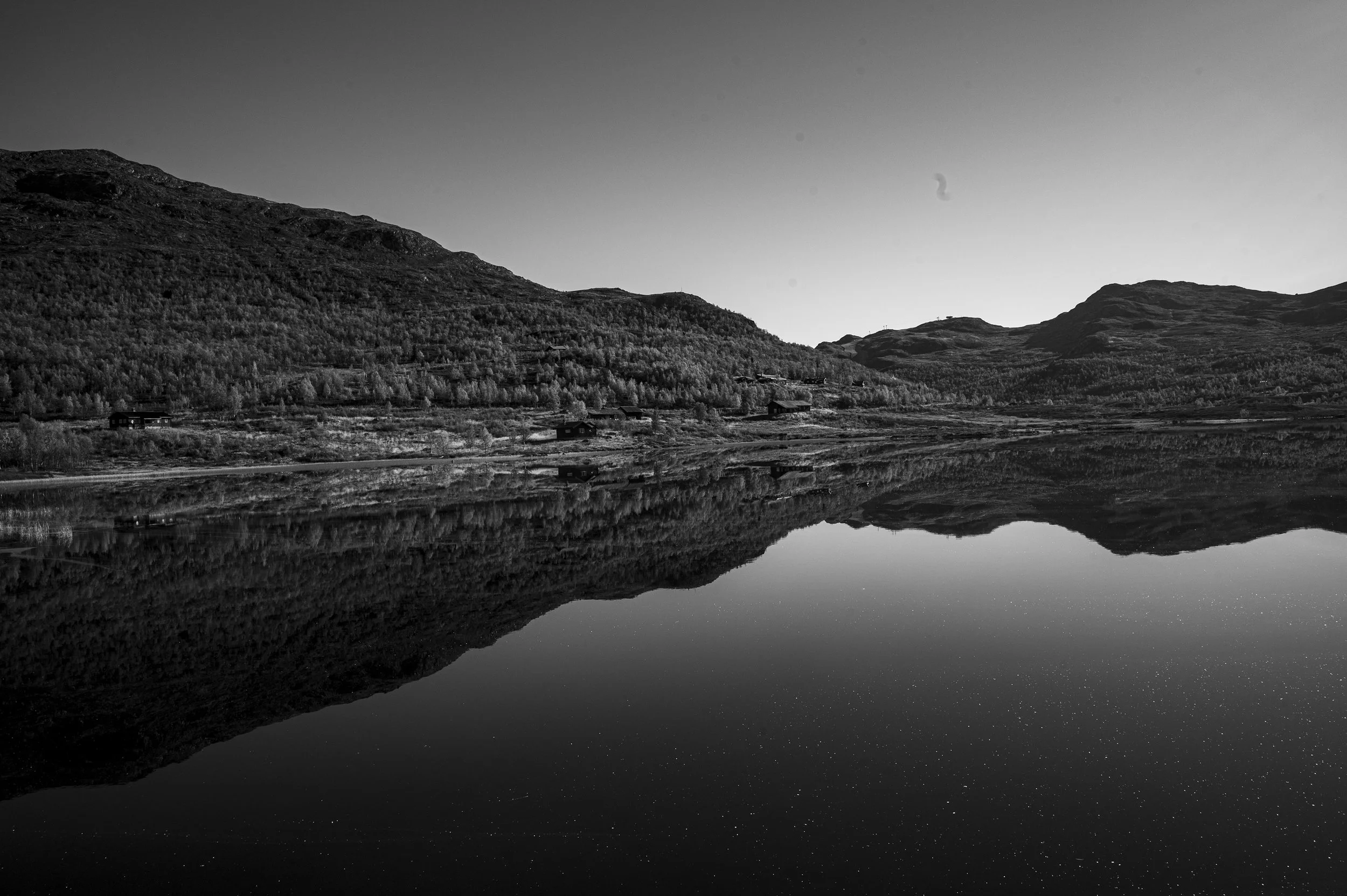 Black and white photo of a calm lake with a mountain range in the background, reflecting the sky and mountains. Small buildings scattered along the shoreline of the lake.