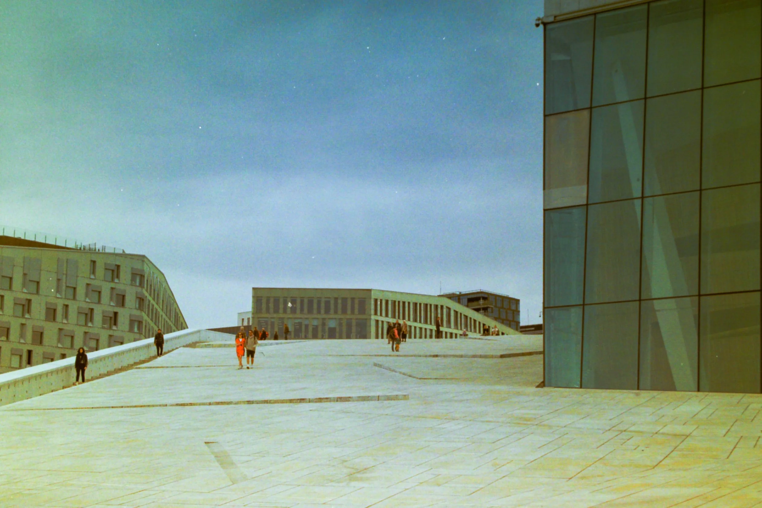 People walking on a modern, curved architectural terrace with glass buildings and a cloudy sky.