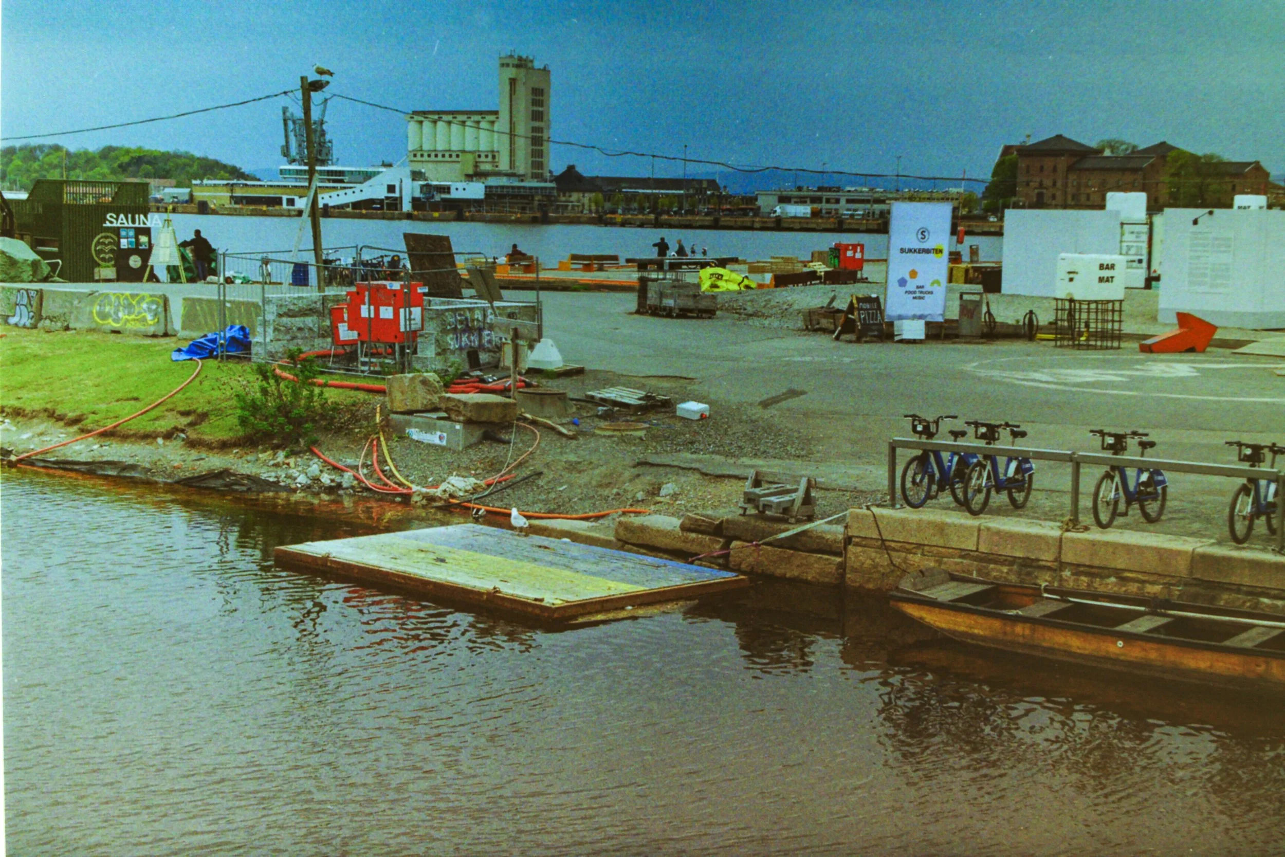 A riverfront scene with construction equipment, bicycles, and a boat. In the background, there are industrial buildings and a bridge over the water.