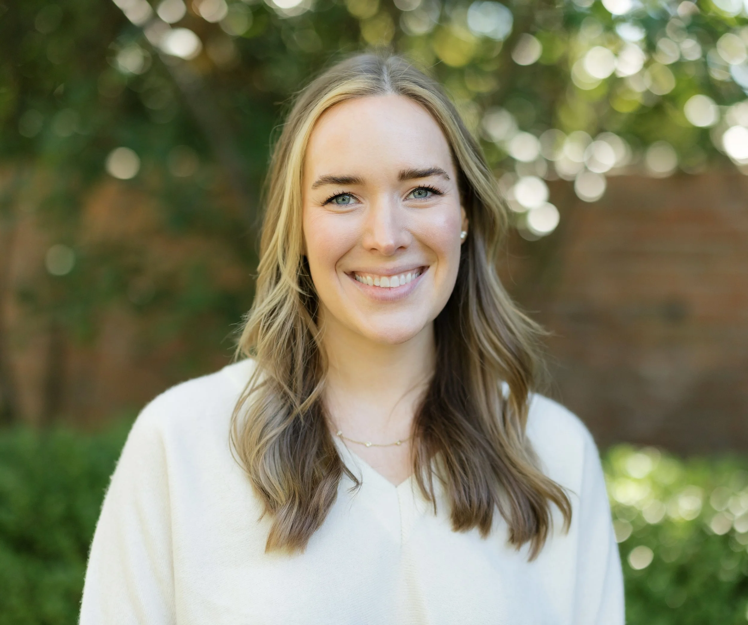 A woman with blonde hair and blue eyes smiling outdoors with trees and a brick wall in the background.