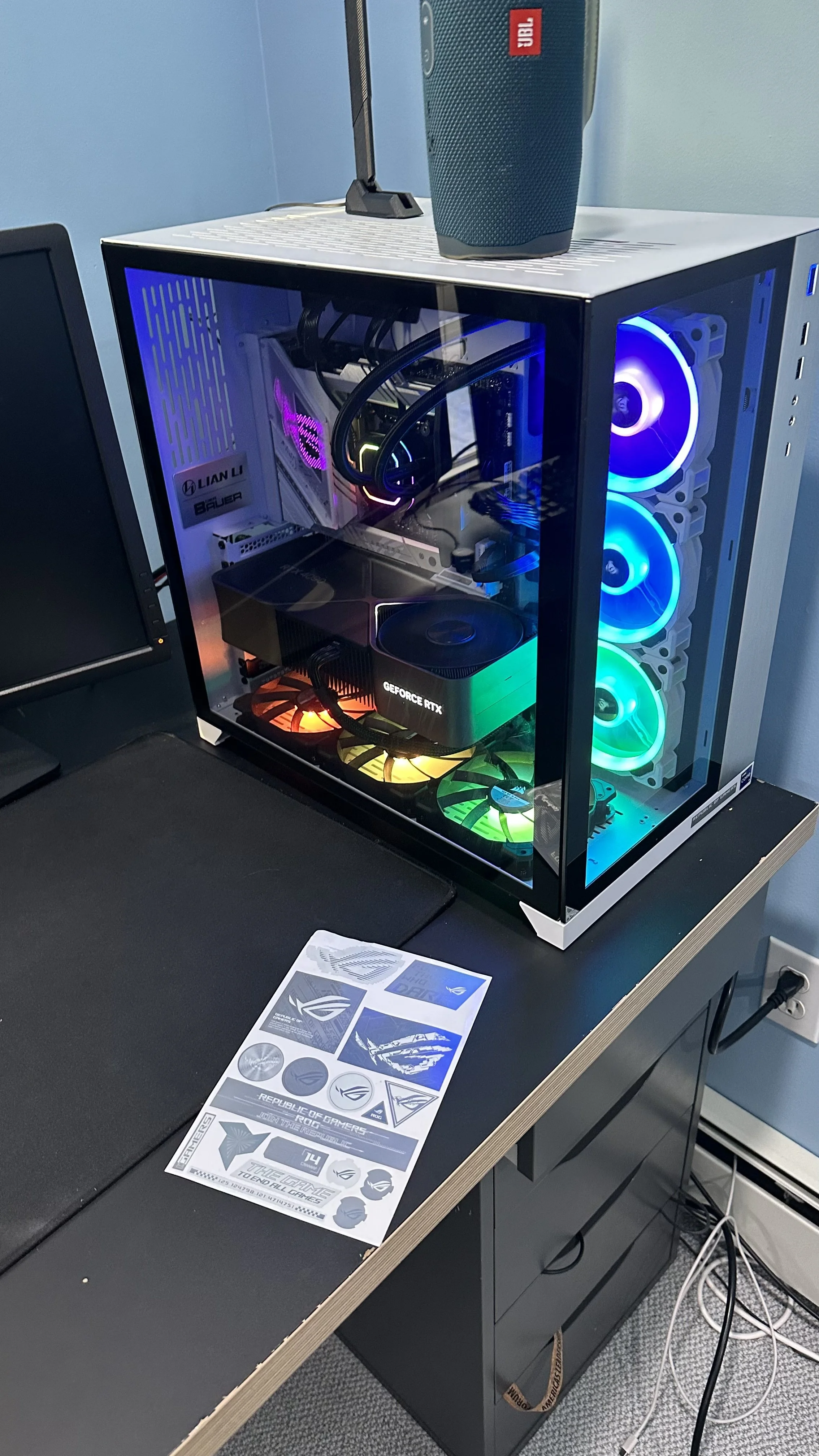 A gaming desktop PC with a transparent side panel displaying internal components, including a GeForce RTX graphics card, colorful RGB cooling fans, and a motherboard. The PC case has a metal top with a JBL speaker placed on it. The setup is on a black desk with a monitor and printed materials nearby.