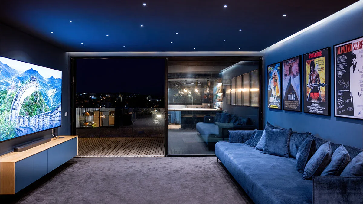 Modern living room at night with a large TV, a sliding glass door leading to a balcony, and framed posters on the wall.