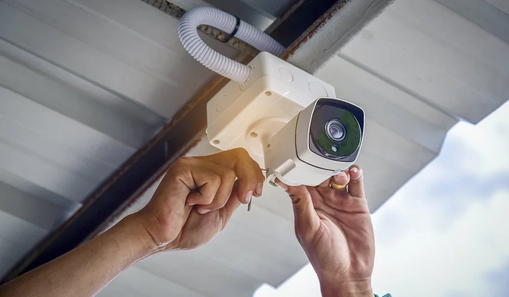 A person installing or maintaining a security camera on an outdoor structure, with hands adjusting the camera under a metal roof or ceiling.