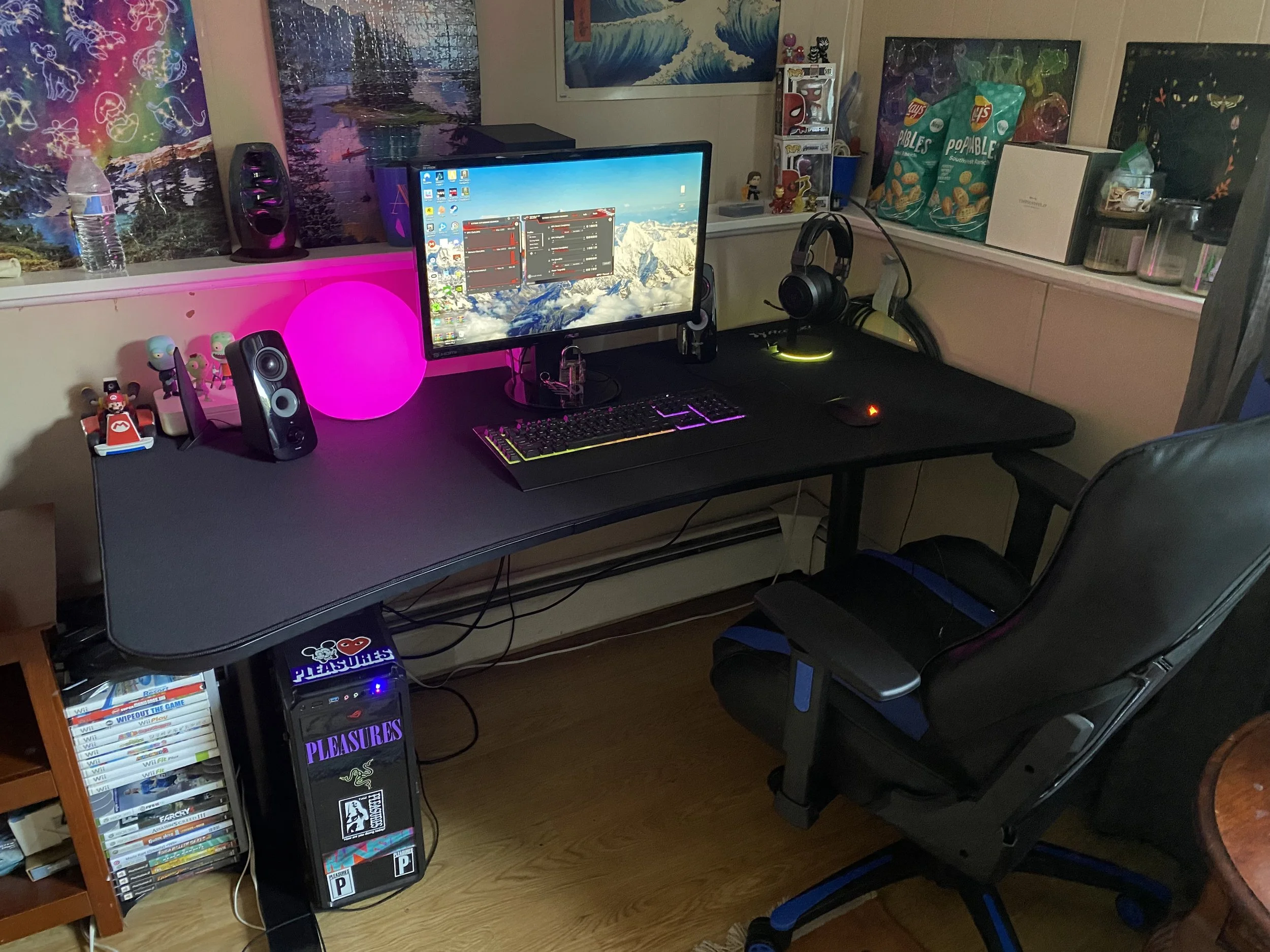 A gaming desk setup with a widescreen monitor, wired keyboard, gaming mouse, and over-ear headphones. The desk also features speakers, a lit pink lamp, and various decorations along the back wall, including posters and snack boxes.
