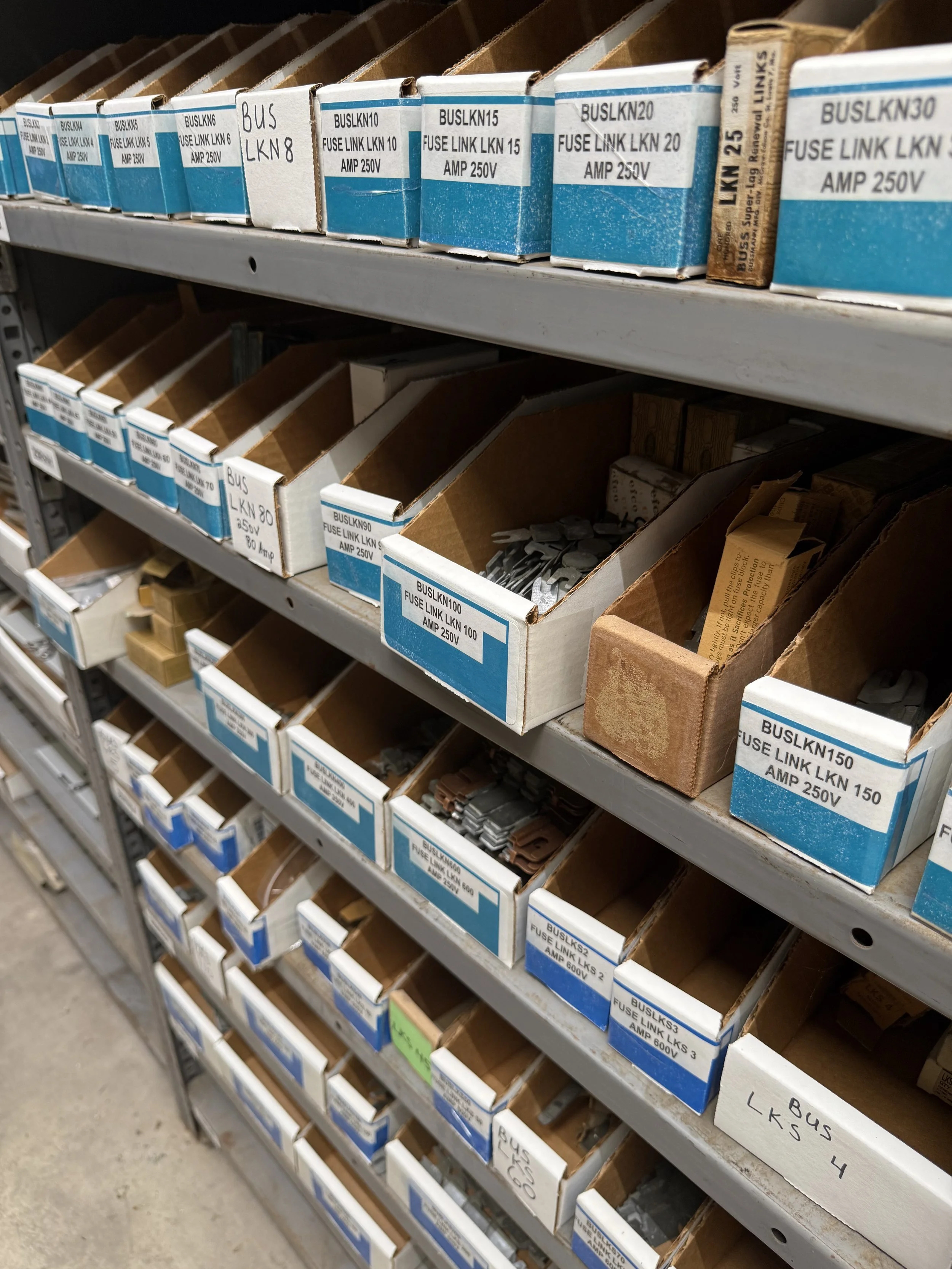 Multiple labeled boxes of electrical fuses and fuse links stored on metal shelves in a hardware store.