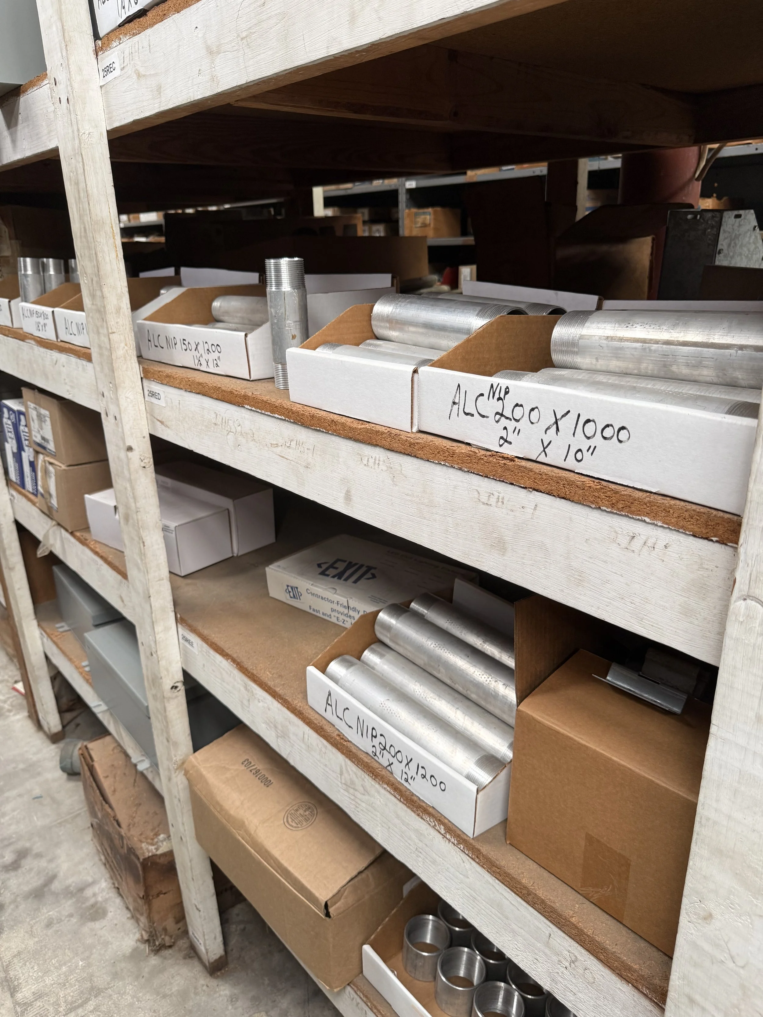 Metal pipes and fittings stored on wooden shelving in a warehouse, labeled with specifications.
