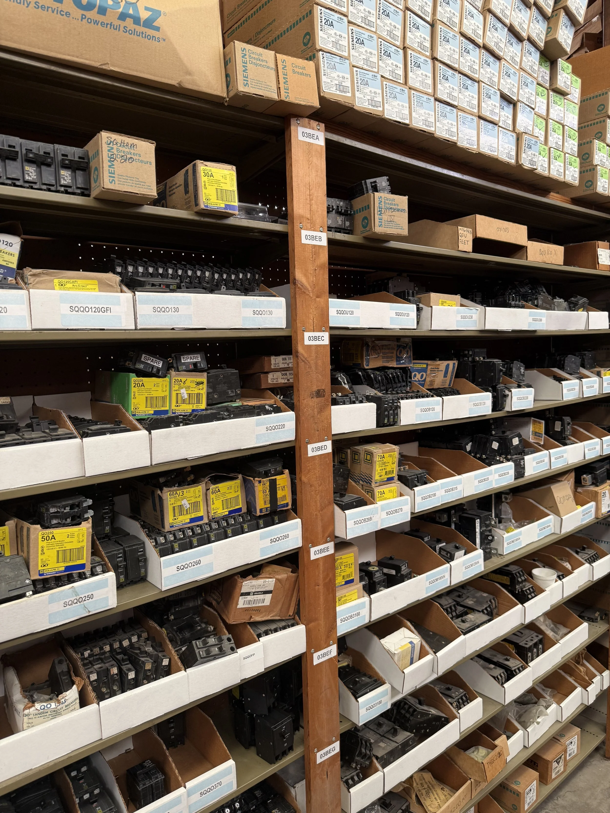 Warehouse aisle with shelves stocked with electrical circuit breakers and fuses, organized in white bins and cardboard boxes.