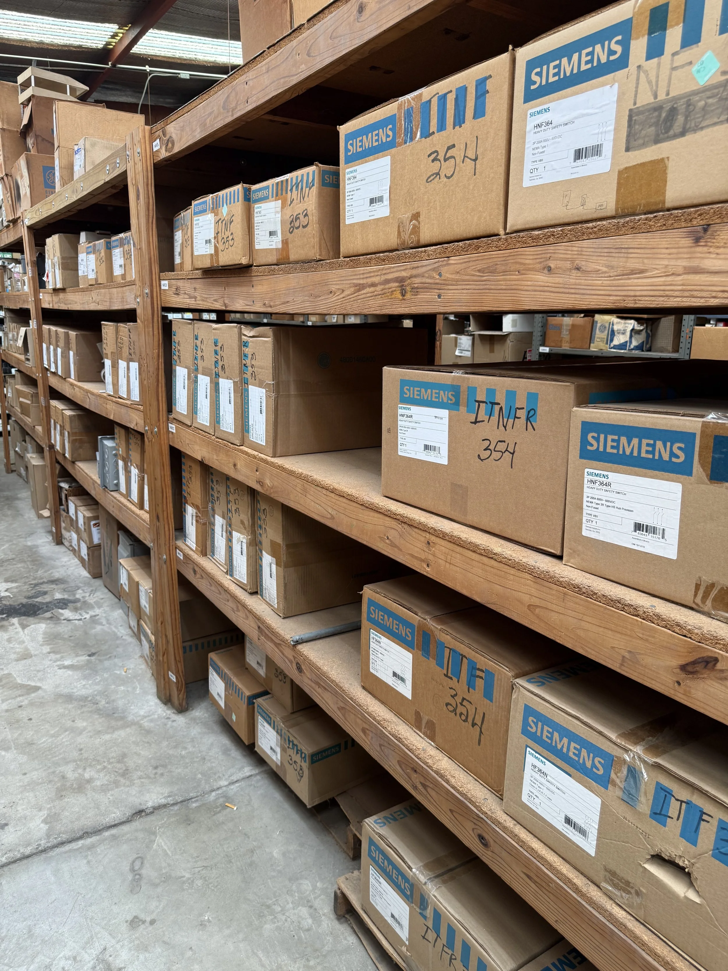 Warehouse shelving with stacked cardboard boxes containing electrical components such as switches, disconnects and load centers.