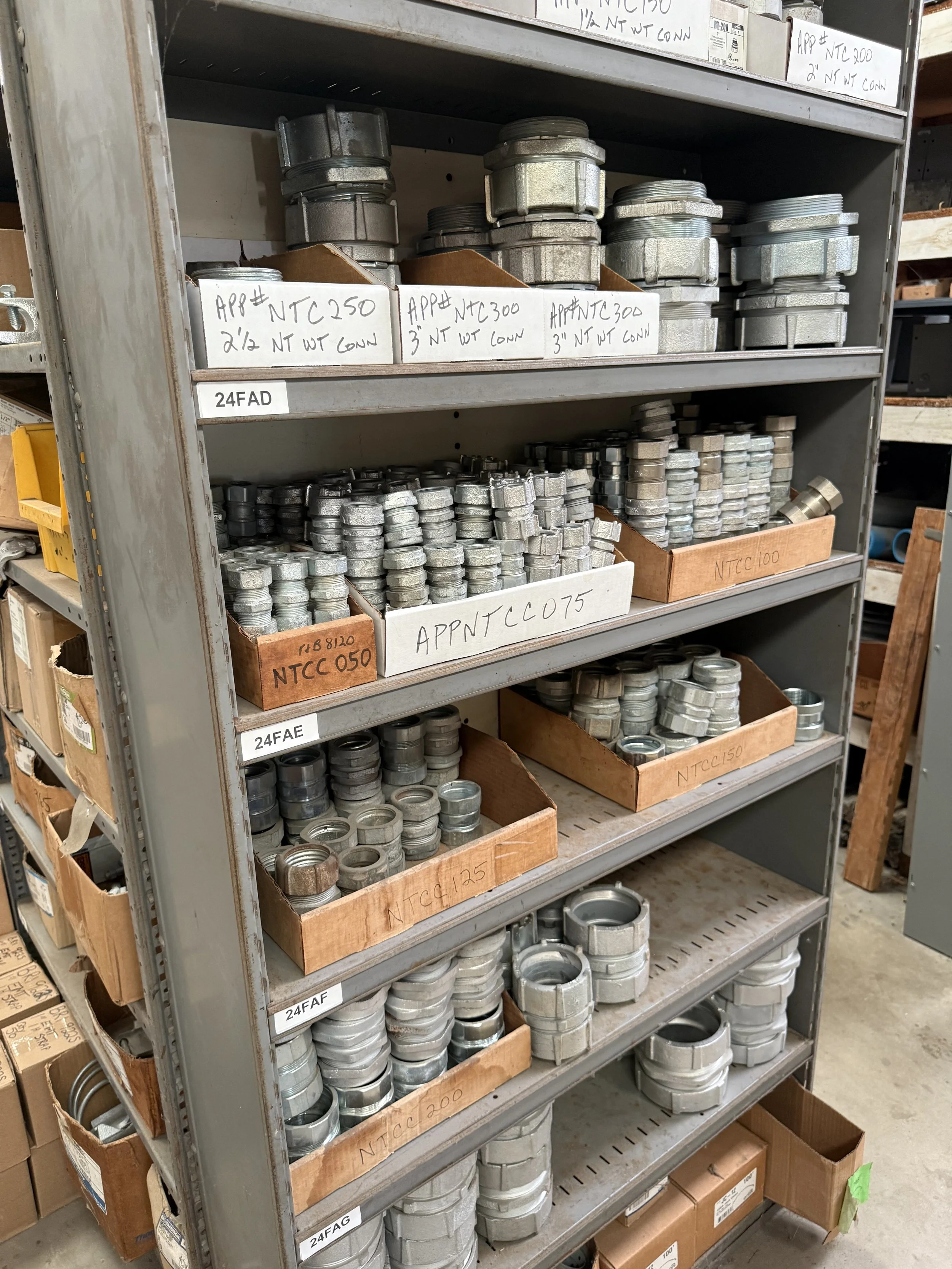 A metal shelving unit filled with metal pipe fittings, water tight couplings, EMT.