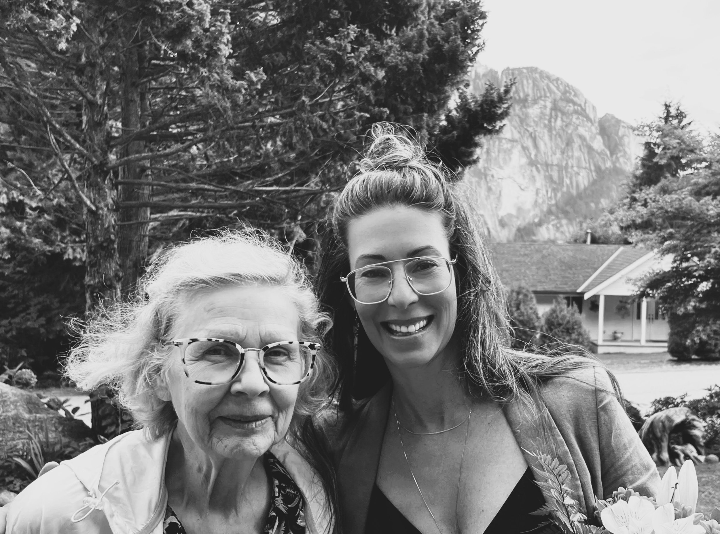 Black and white photo of two women outdoors, one elderly with glasses and white hair, the other younger with long wavy hair and glasses, smiling at the camera.Helping a senior parent downsize.
