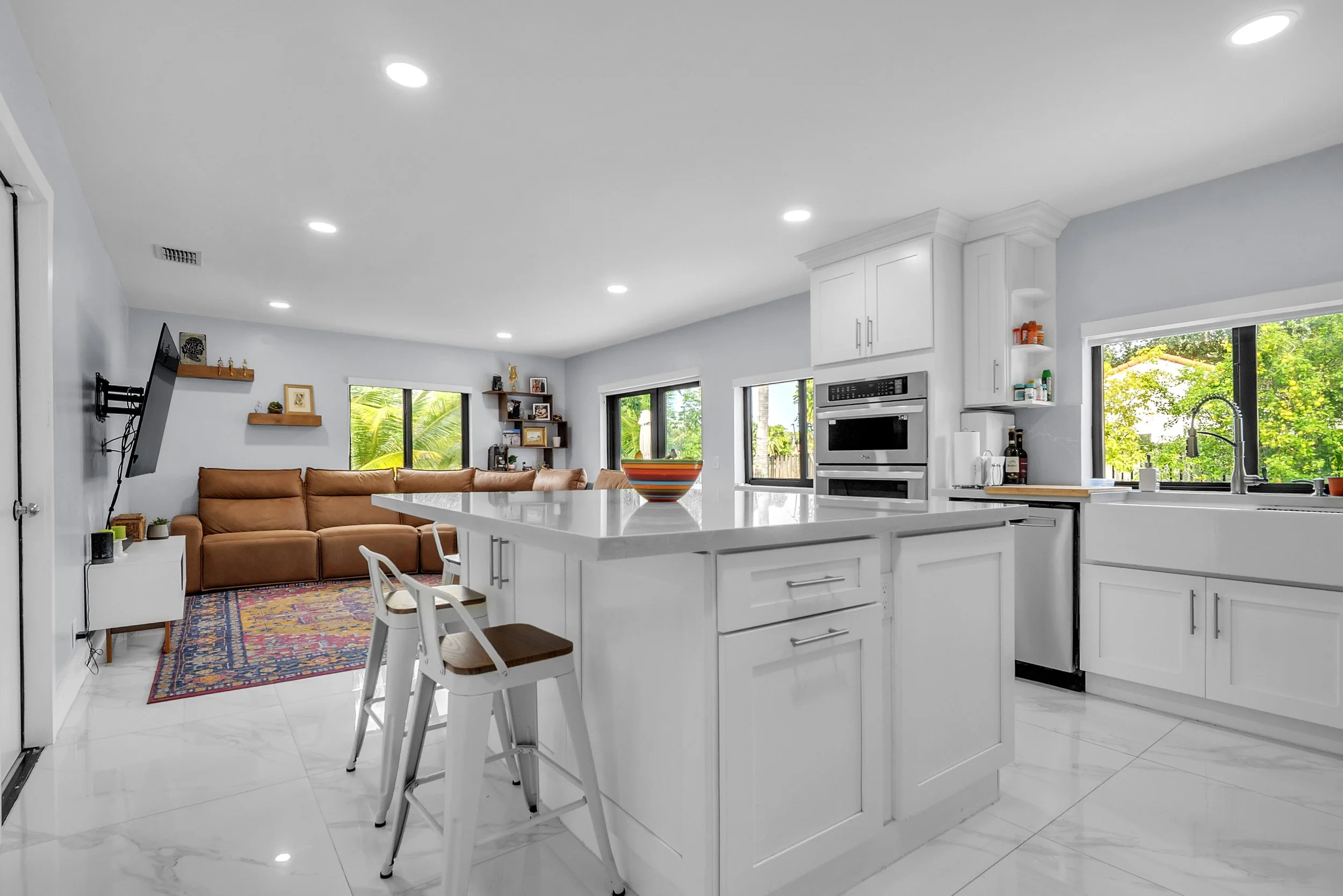 Open-plan kitchen and living room with white cabinetry, marble flooring, a central island with a bowl, window overlooking greenery, brown leather sofa, TV on wall, and natural light.
