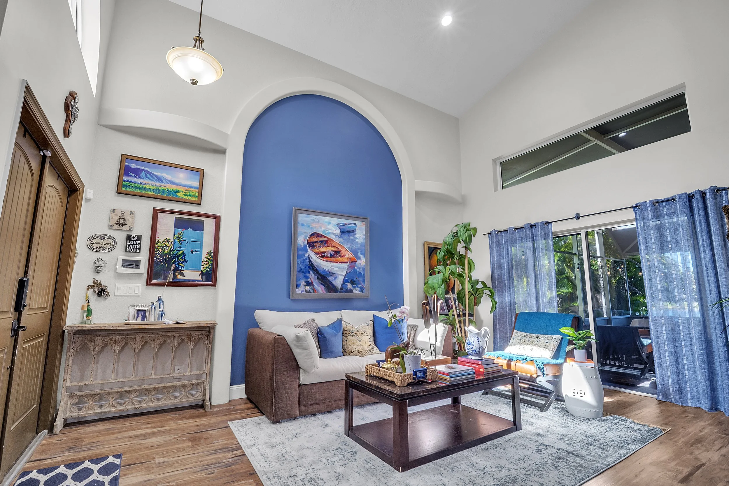 Living room with white walls, blue accent wall, wooden floor, and large window with blue curtains. Features a beige sofa with pillows, a dark wooden coffee table with books and plants, a blue lounge chair, and wall art, including paintings and decorations.