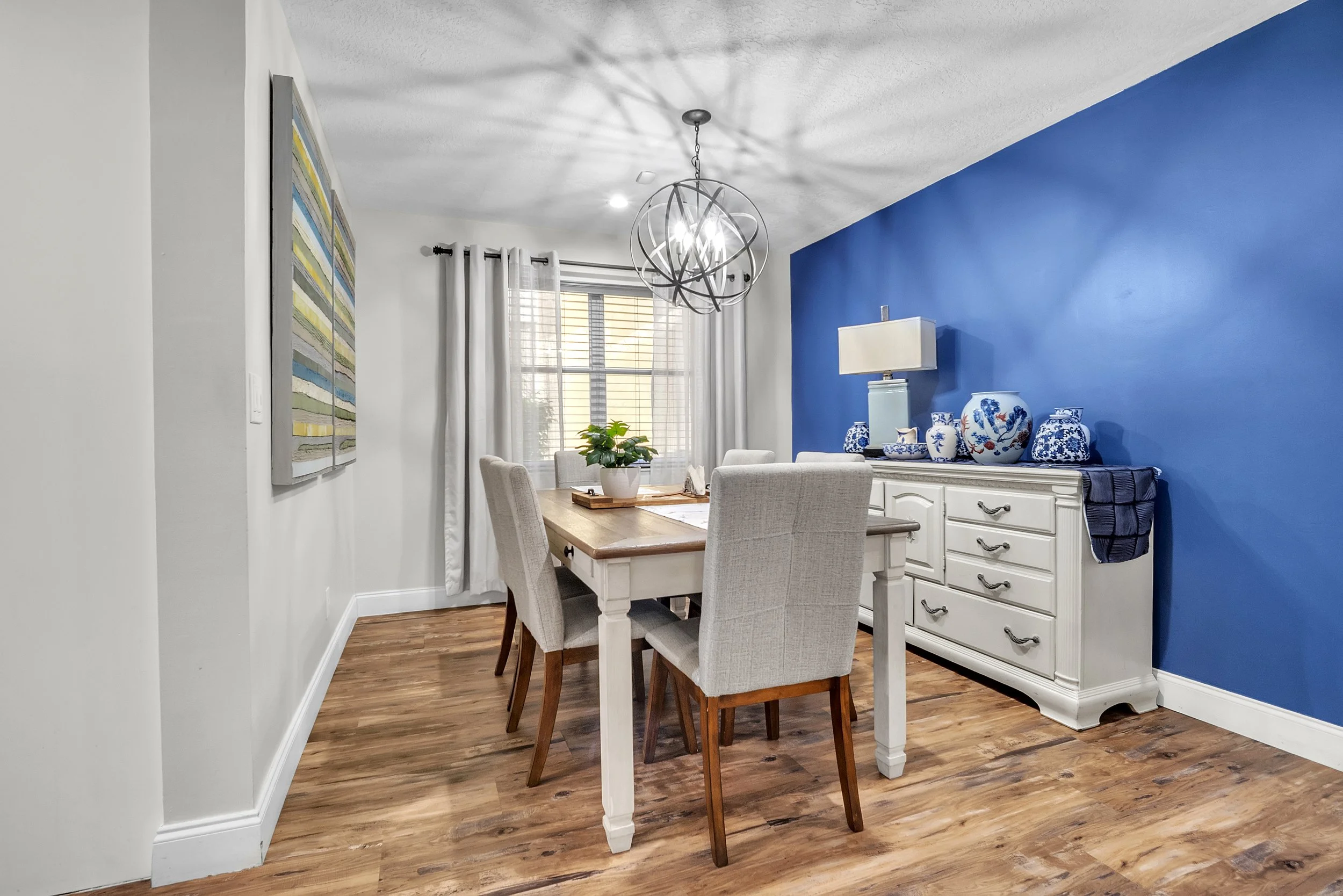 Dining room with a wooden table, six upholstered chairs, a sideboard with blue and white decorative vases, a lamp, and a blue accent wall. White curtains and a window are in the background. Wooden flooring and a modern chandelier are overhead.