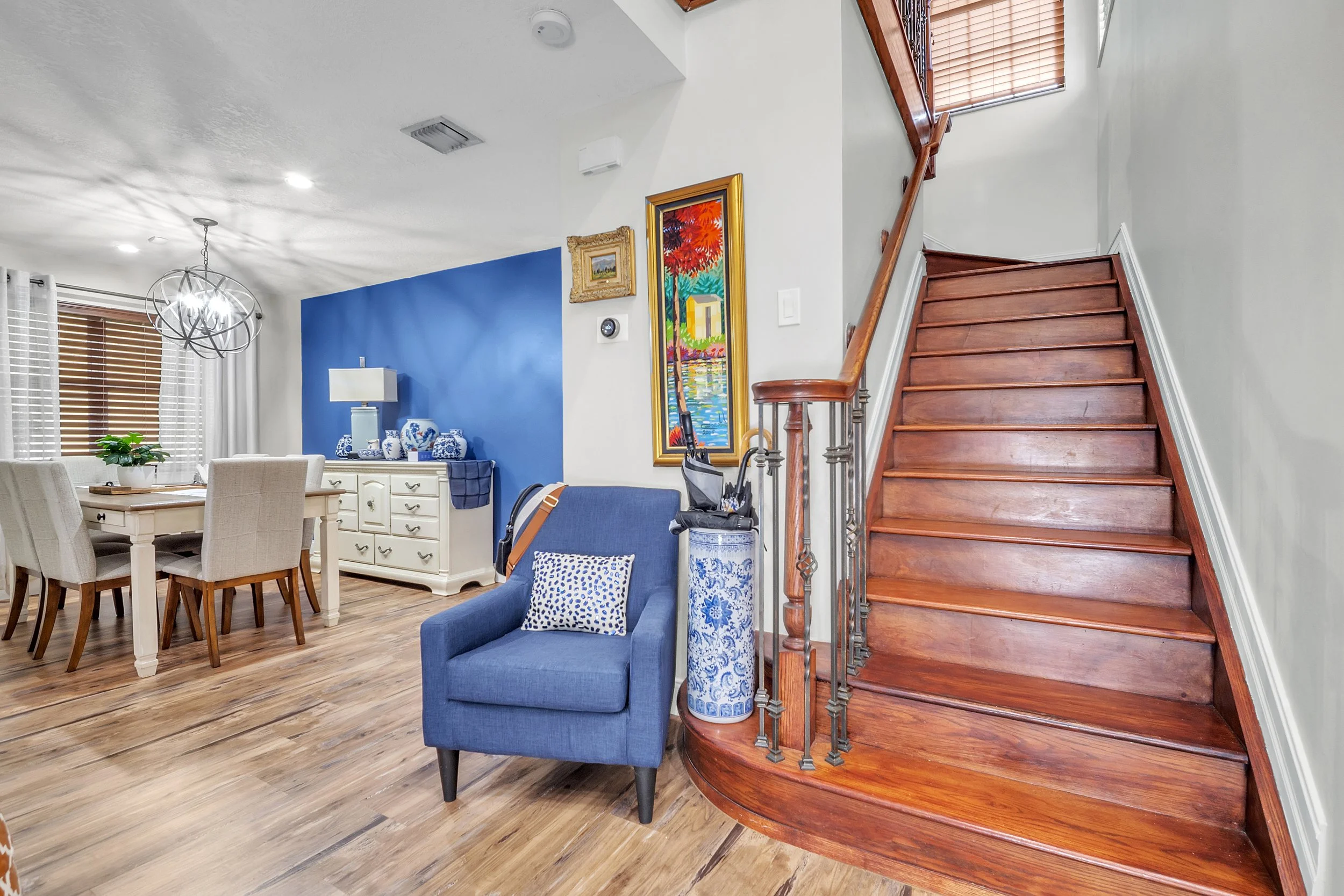 Interior view of a living room with wooden floors, a blue accent wall, a blue armchair with a patterned pillow, a white sideboard with decorative blue and white pottery, a dining table with cream-colored chairs, a modern chandelier, and a wooden staircase with a metal railing.