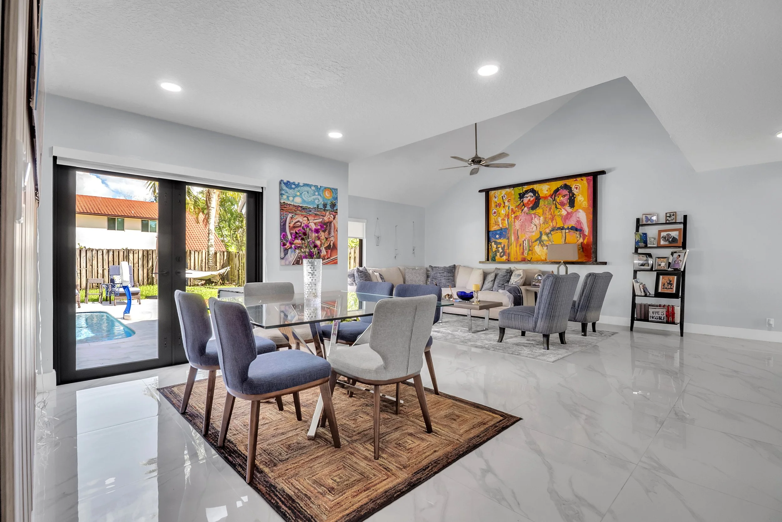 Modern living room with glass dining table, eight chairs, artwork on white walls, large window showing backyard with pool, and a cozy seating area with a white sofa and armchairs.